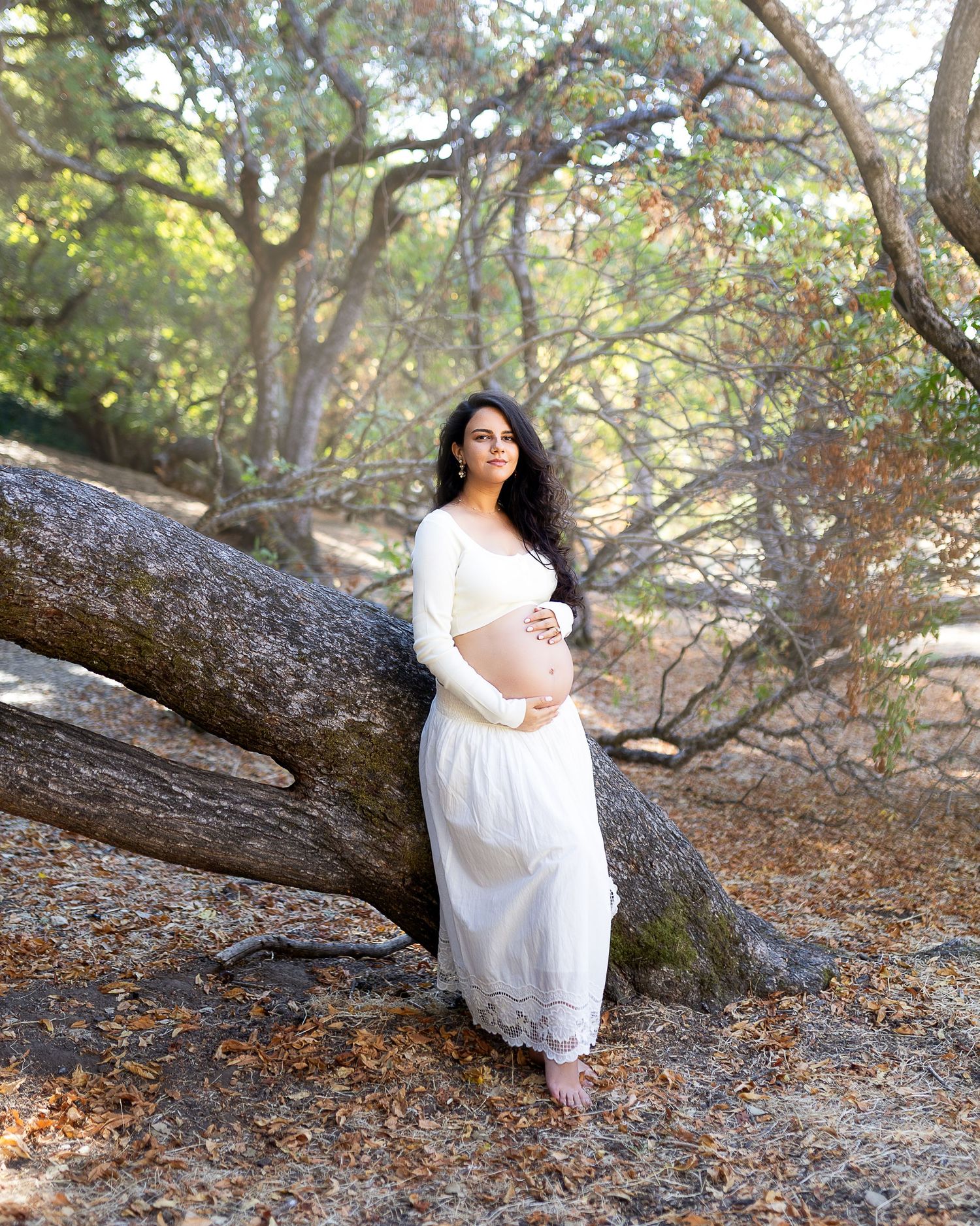 A person in a flowing white dress poses gracefully against fallen autumn leaves and tree branches in a forest setting.