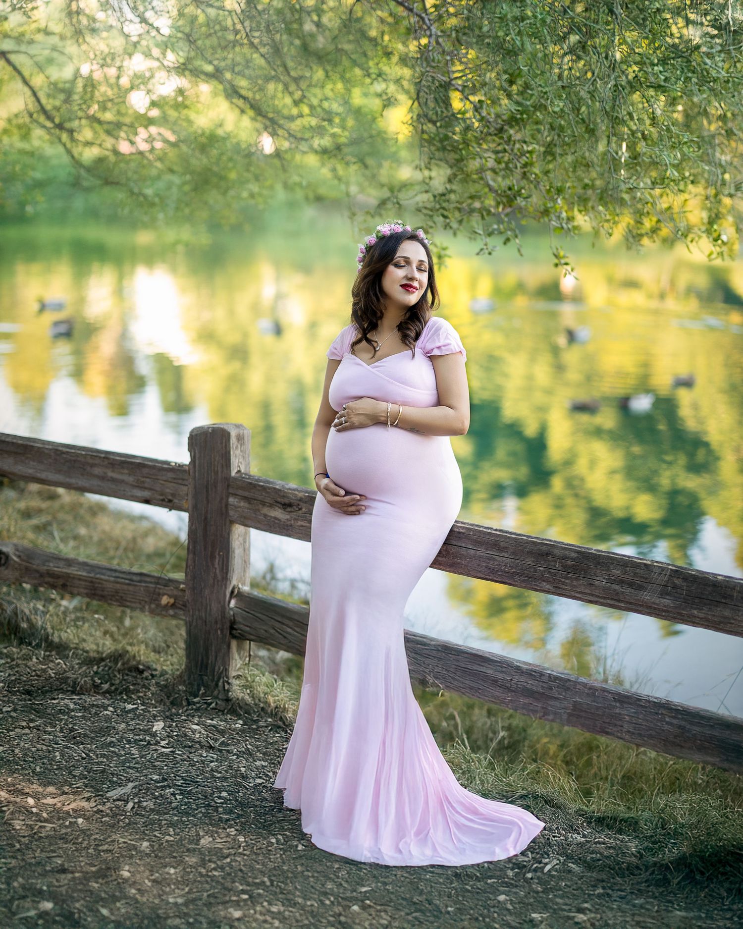 A woman in a flowing pink dress stands by a rustic wooden fence near a tranquil pond during golden hour.