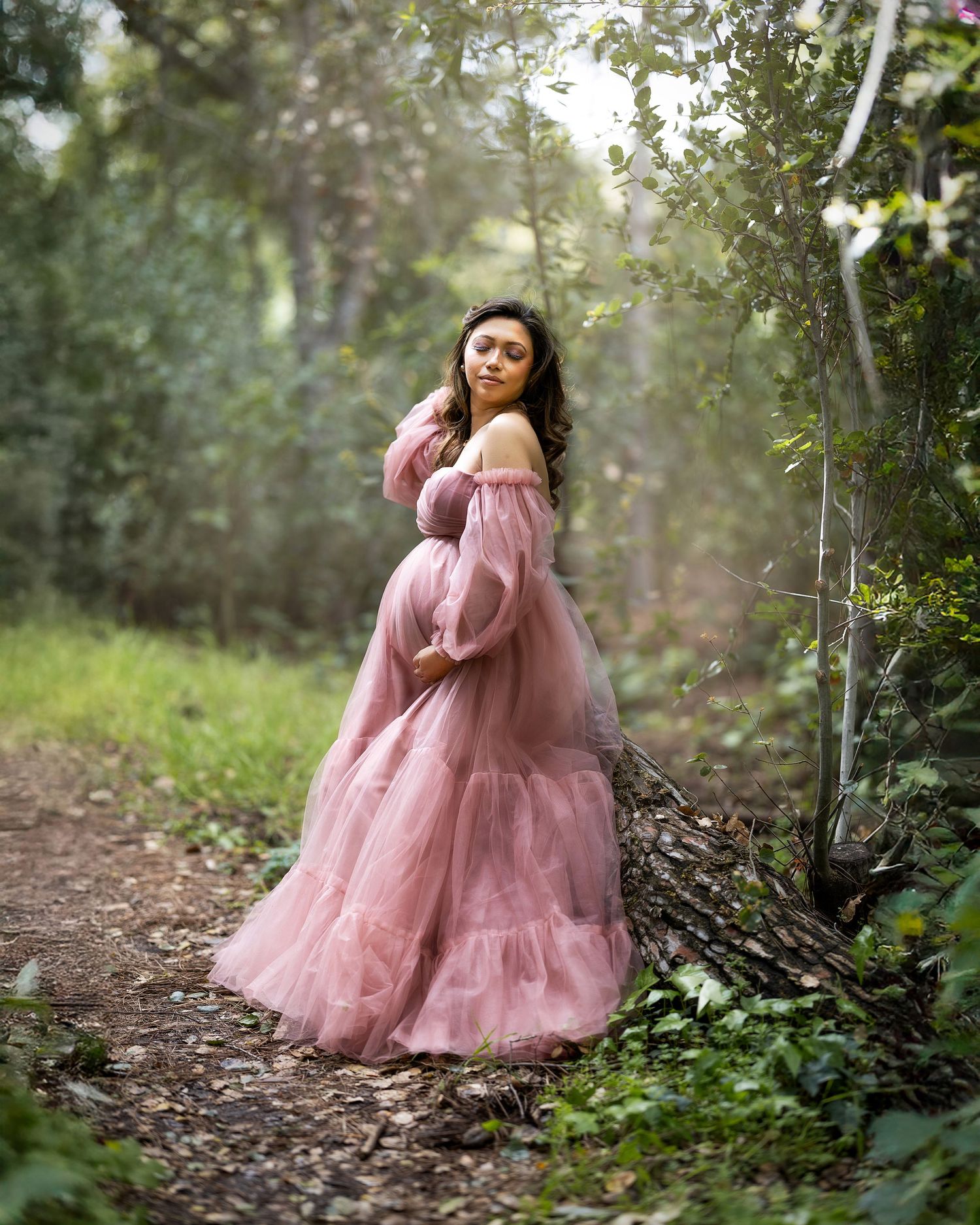 A dreamy maternity photo shows an expectant mother in a flowing pink gown standing on a forest path in soft lighting.