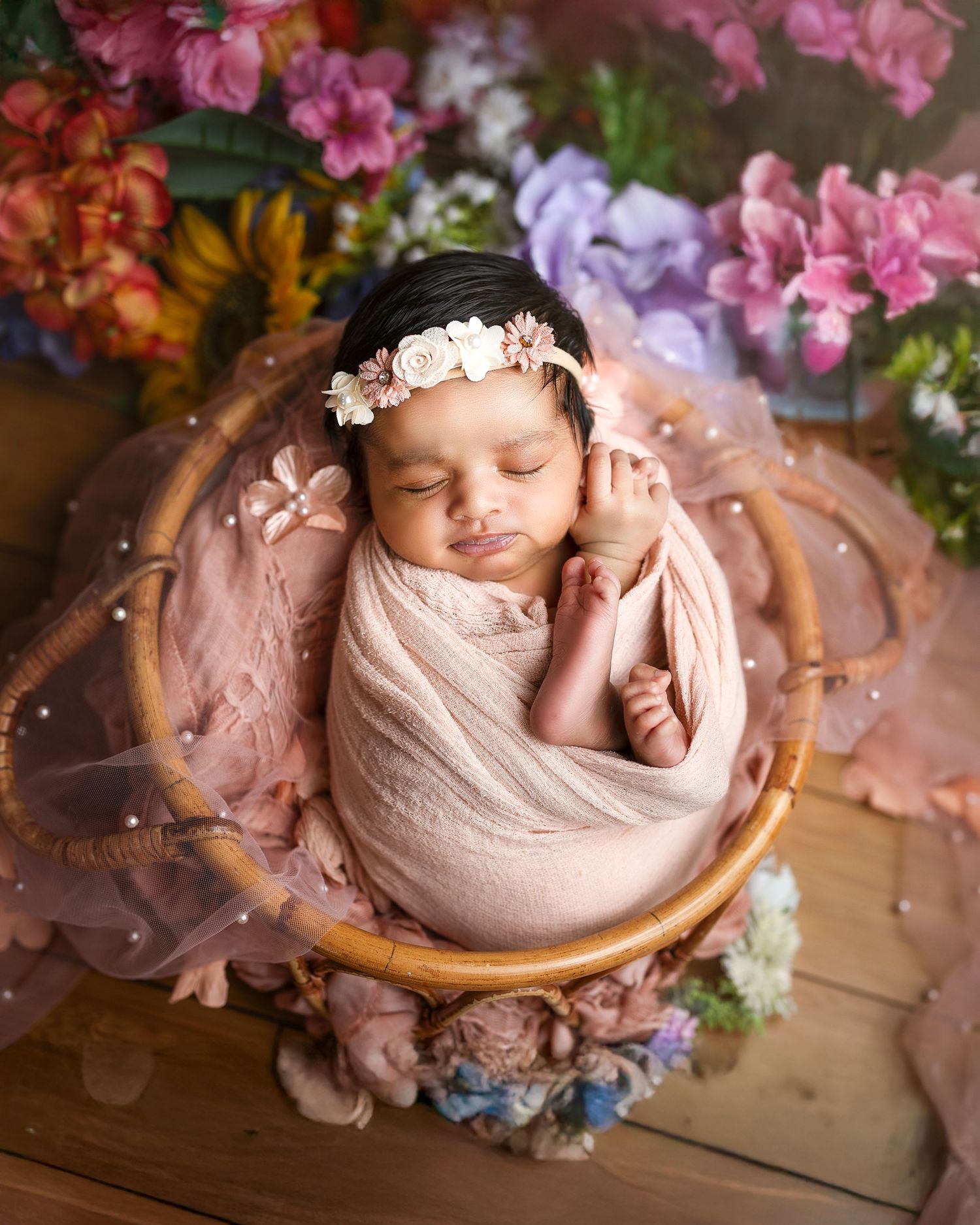 Sleeping newborn wrapped in pink swaddle rests in wooden bowl surrounded by colorful spring flowers and tulle fabric.