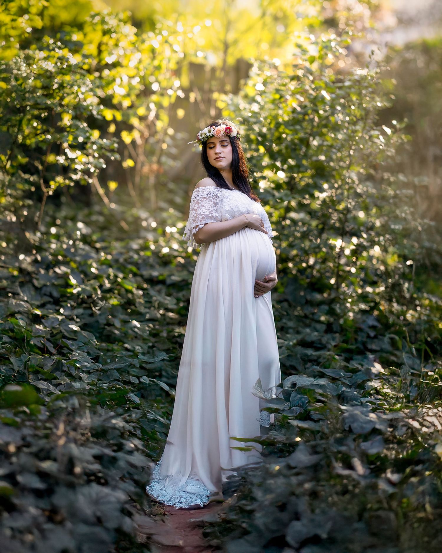 A maternity photoshoot in a dreamy garden setting with soft sunlight filtering through trees and white lace dress details.