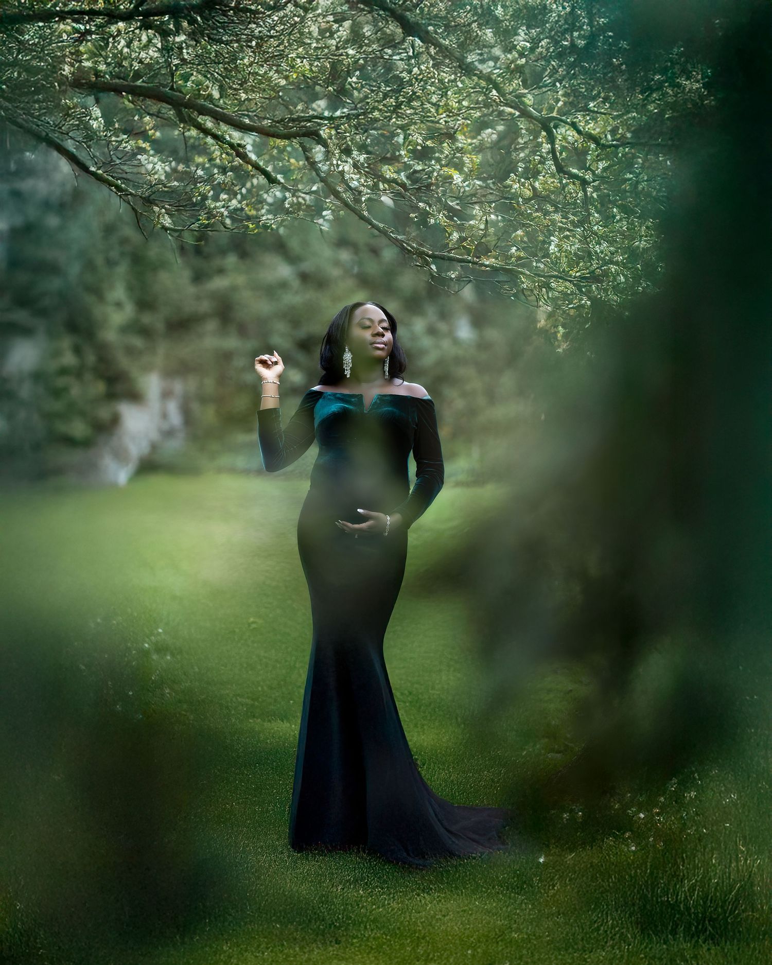 A maternity photo in a mystical forest setting with blossoming spring trees and a flowing navy blue gown.