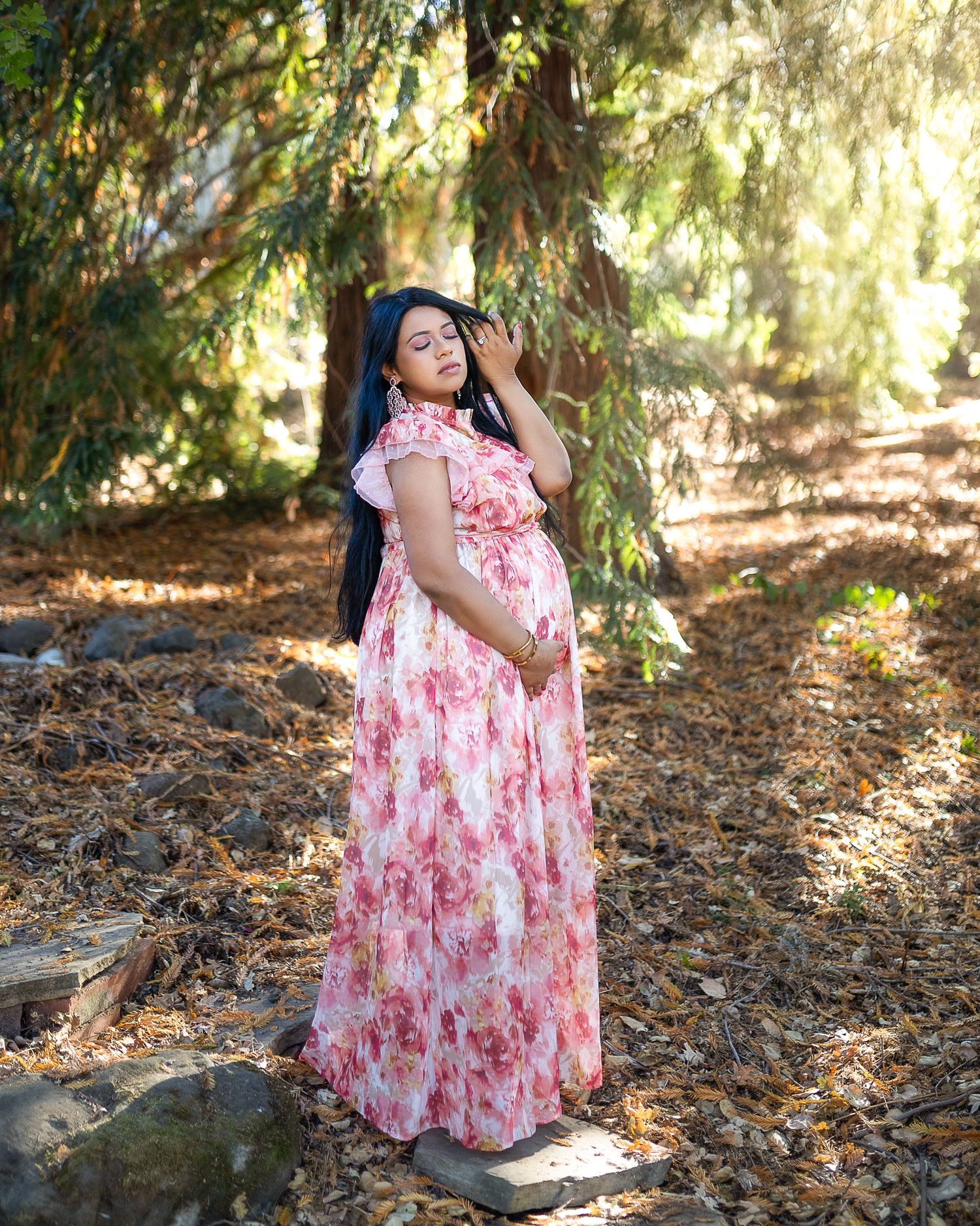 A pregnant woman in a pink floral maxi dress poses outdoors under dappled sunlight and hanging tree branches.
