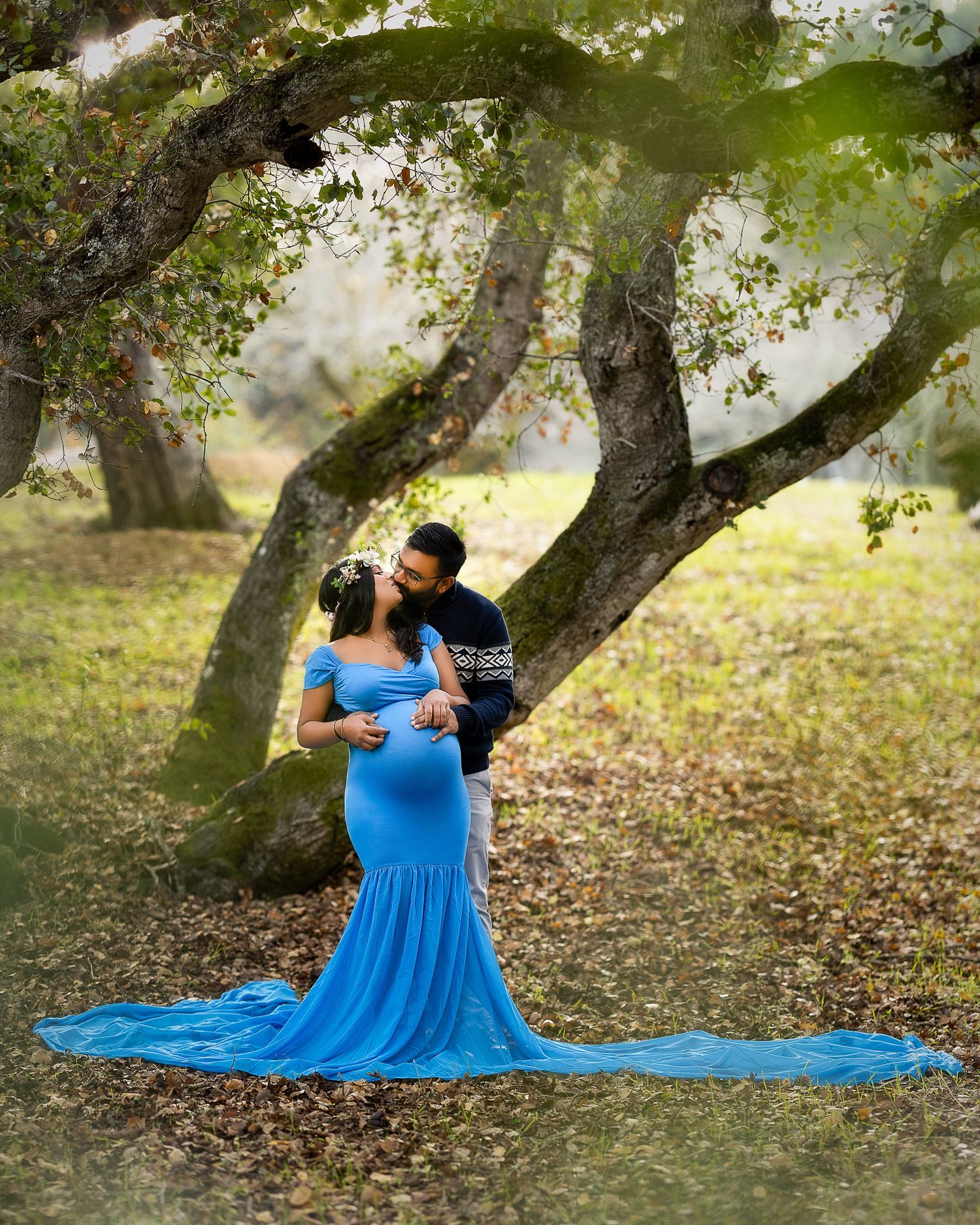 A flowing bright blue maternity gown cascades across fallen leaves beneath oak trees in a natural outdoor setting.