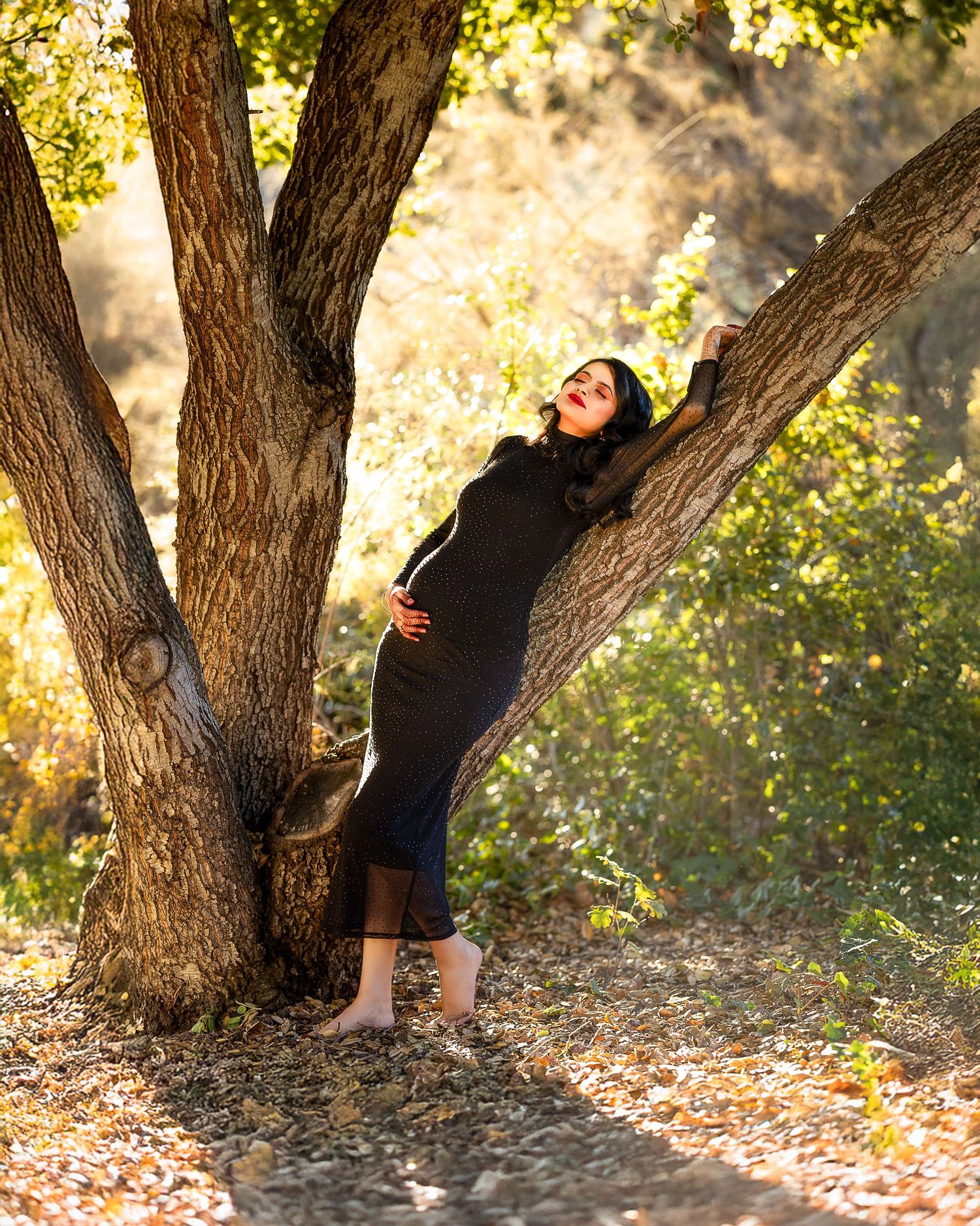 A person in black clothing sits against a tree trunk in dappled sunlight during a golden autumn afternoon.