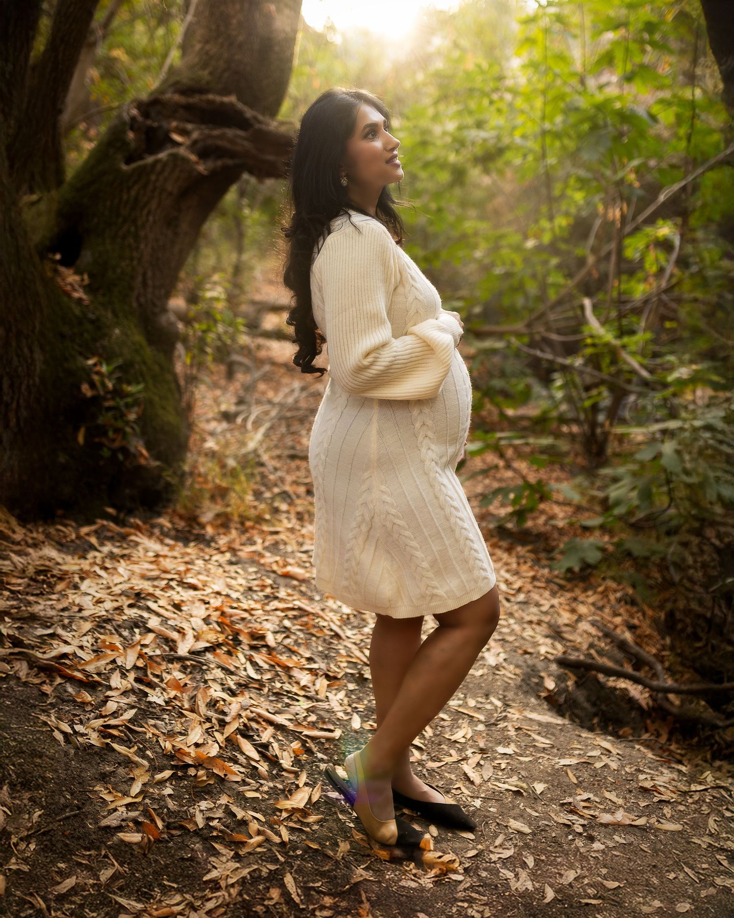 A figure in a flowing white dress twirls through a sunlit forest path covered in fallen autumn leaves.