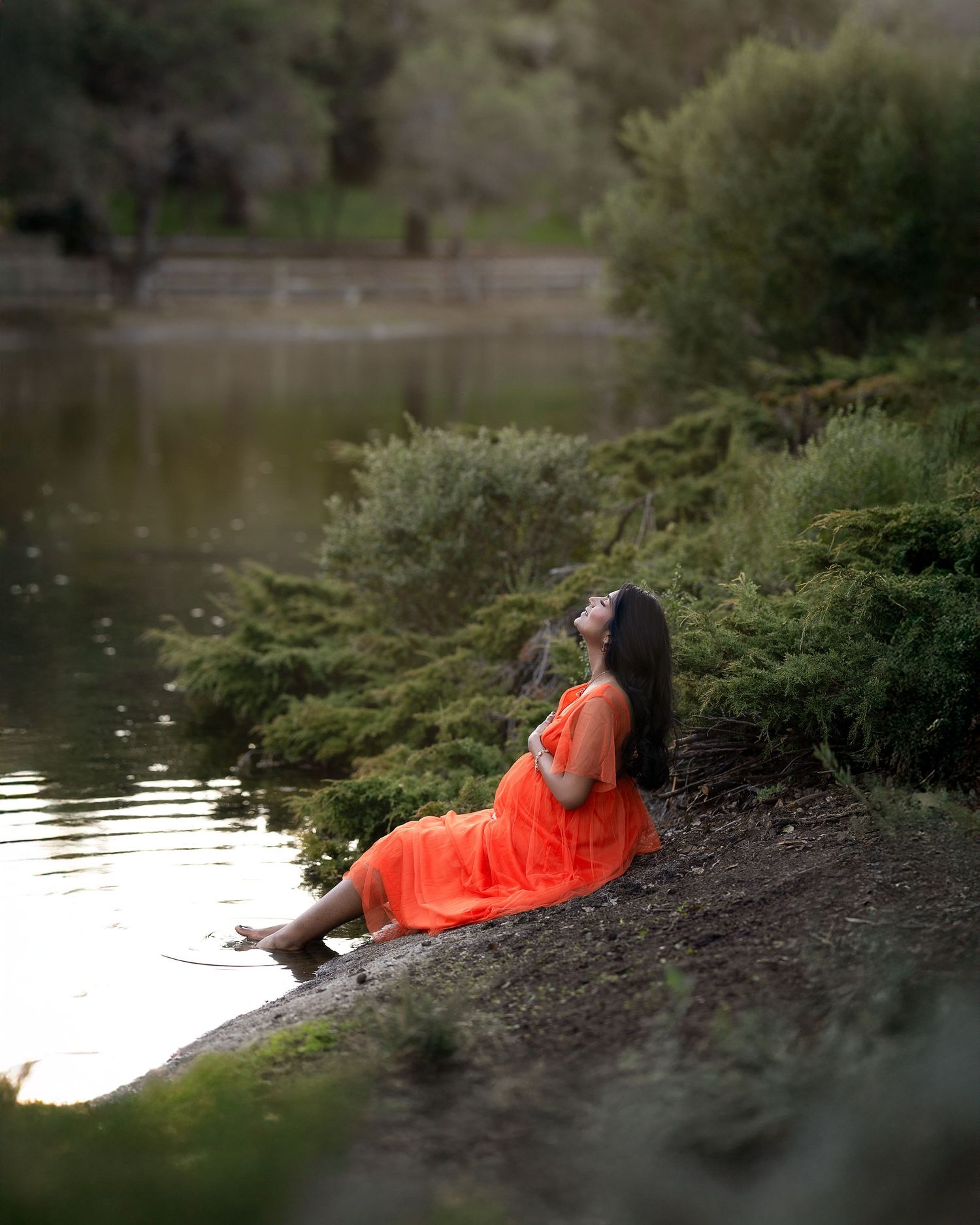 A person in a bright orange dress reclines on the grassy bank of a tranquil lake at sunset.