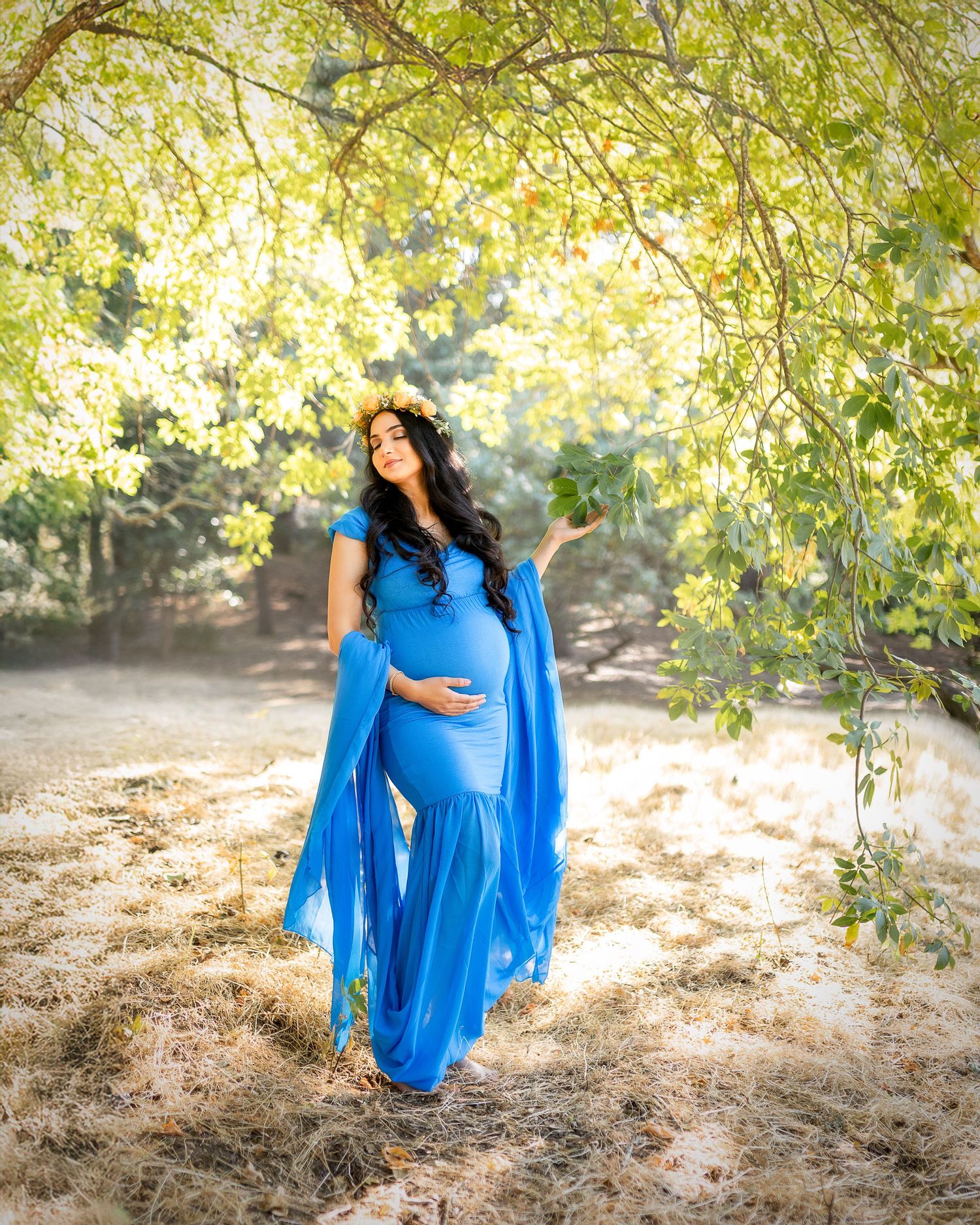 A pregnant woman in a flowing royal blue dress poses under golden autumn trees in a natural outdoor setting.