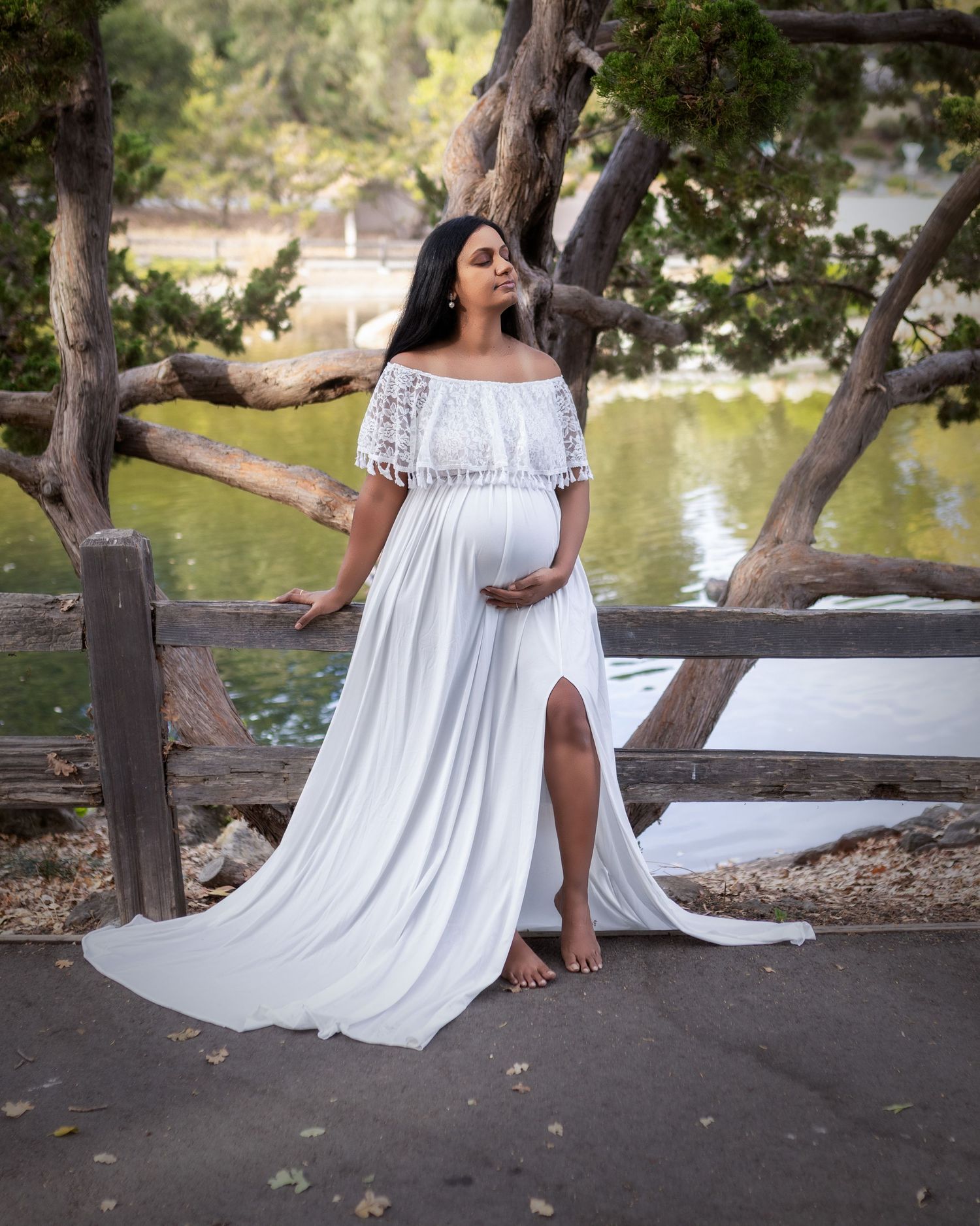 A flowing white off-shoulder maternity dress with lace details photographed in a scenic park setting by a lake.