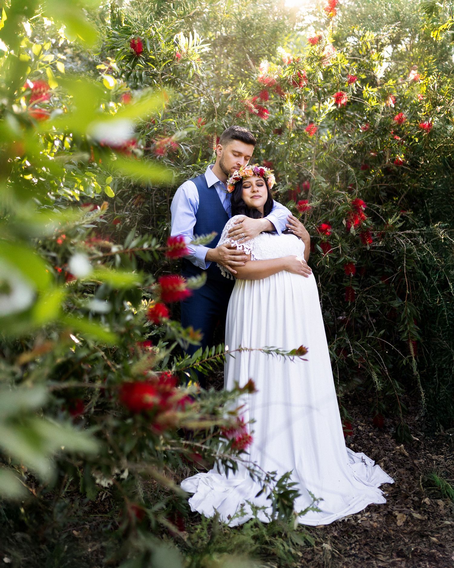 A couple shares a romantic moment in a garden with vibrant red flowers and lush greenery in the background.