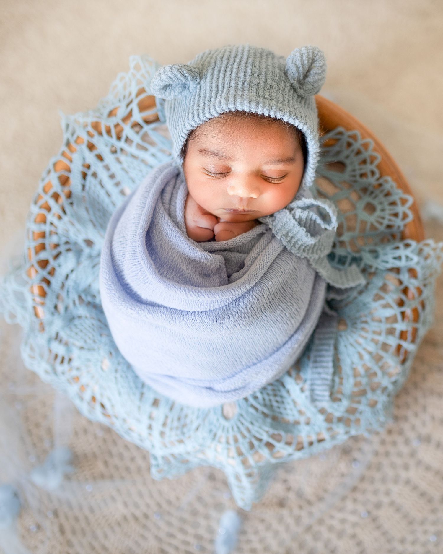 Sleeping newborn wrapped in soft blue knit wear and hat positioned on delicate lace doily.