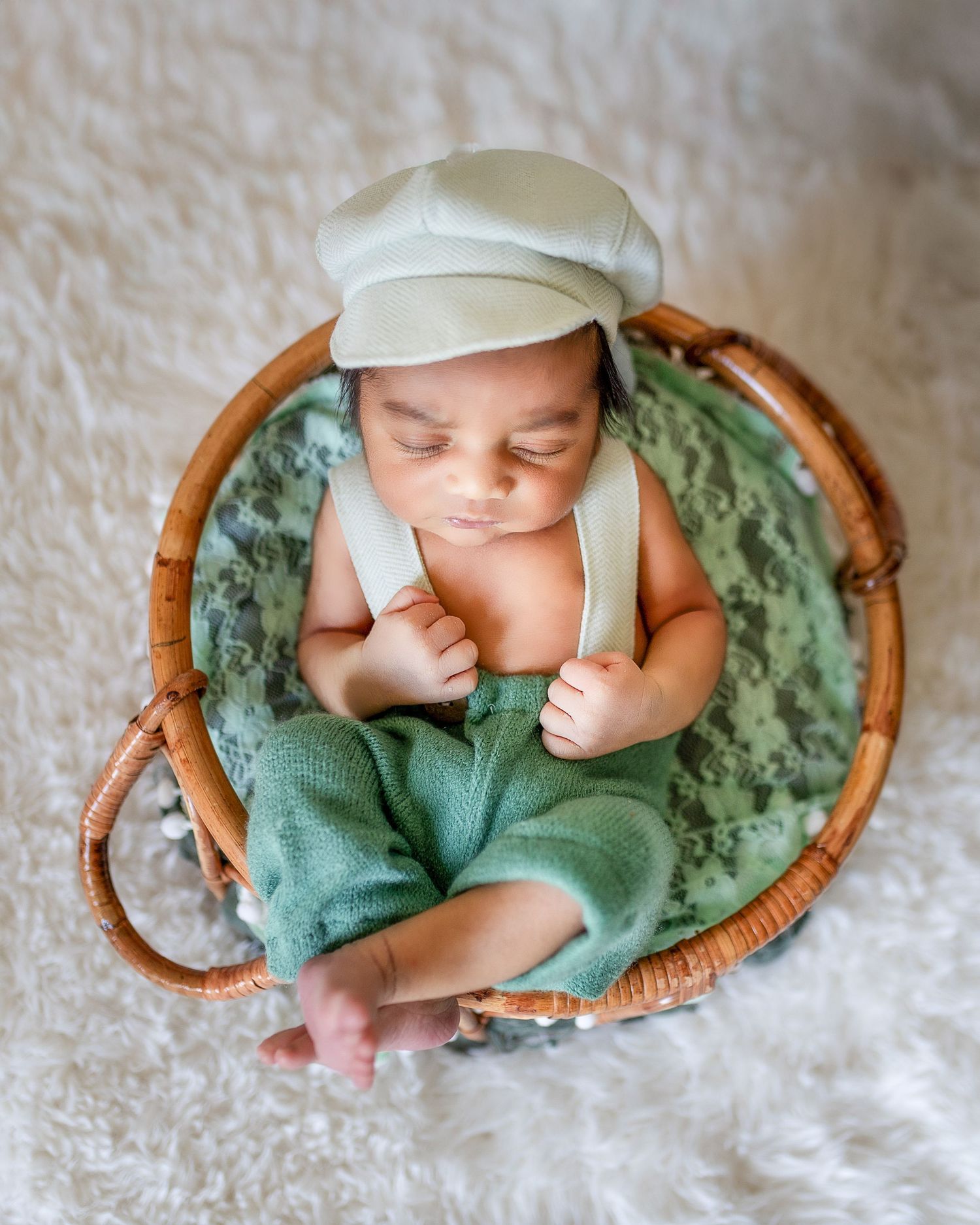 A sleeping newborn in green swaddle rests peacefully in a curved wooden basket on white textured fabric.