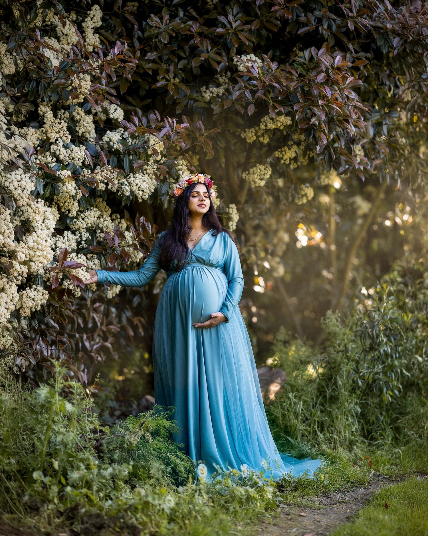 A person in a flowing turquoise dress poses gracefully among blooming white flowers in a garden setting at sunset.