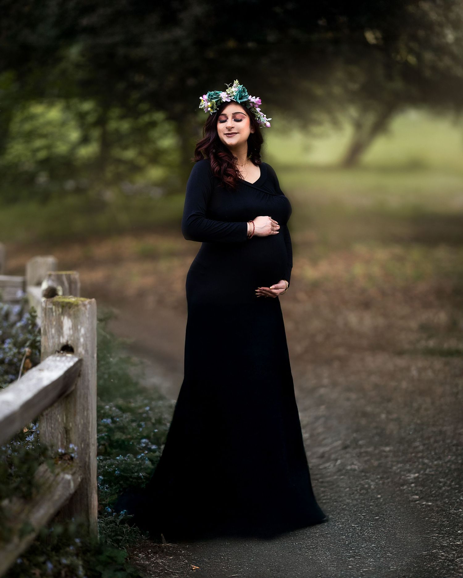 A woman in a flowing black dress stands gracefully on a wooded path during a maternity photoshoot.