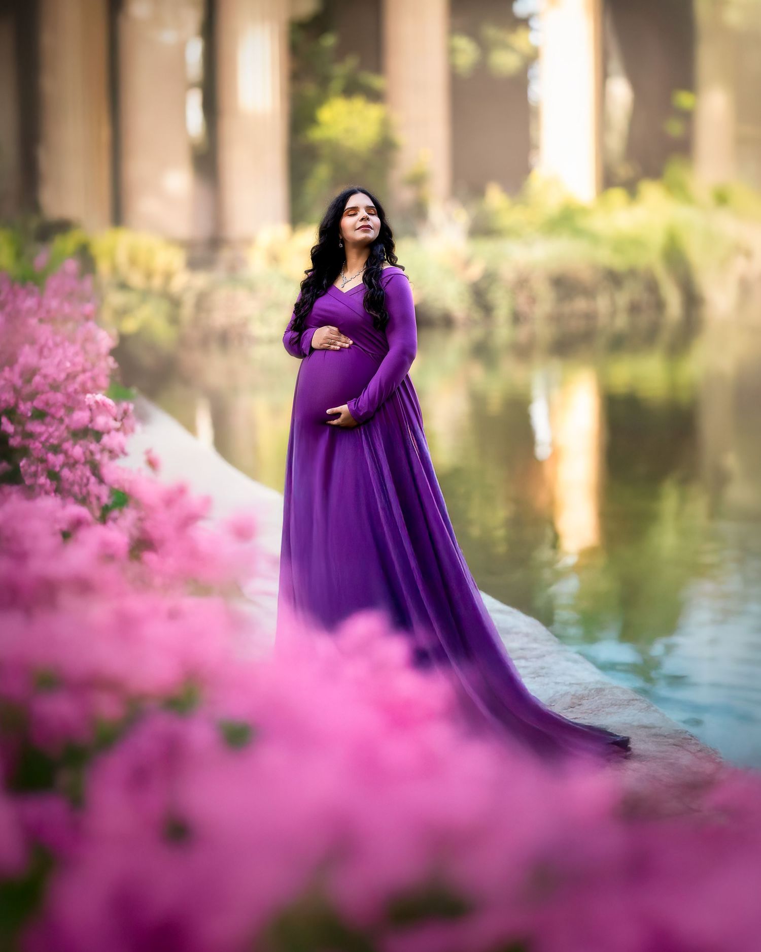 A woman in a flowing purple maternity gown poses elegantly by a lake surrounded by blooming pink flowers.