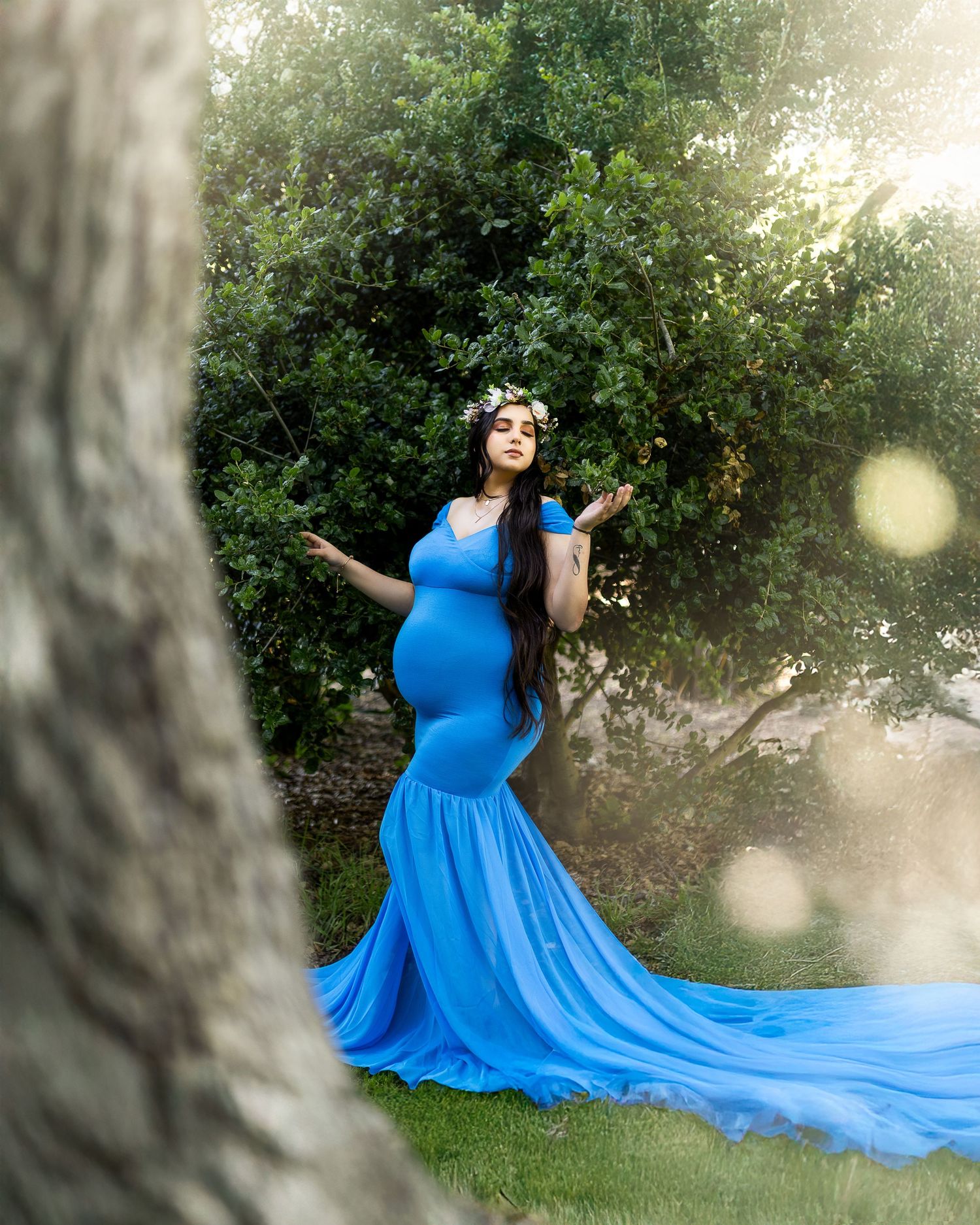 A flowing bright blue maternity gown creates an ethereal effect against a sunlit natural outdoor backdrop.