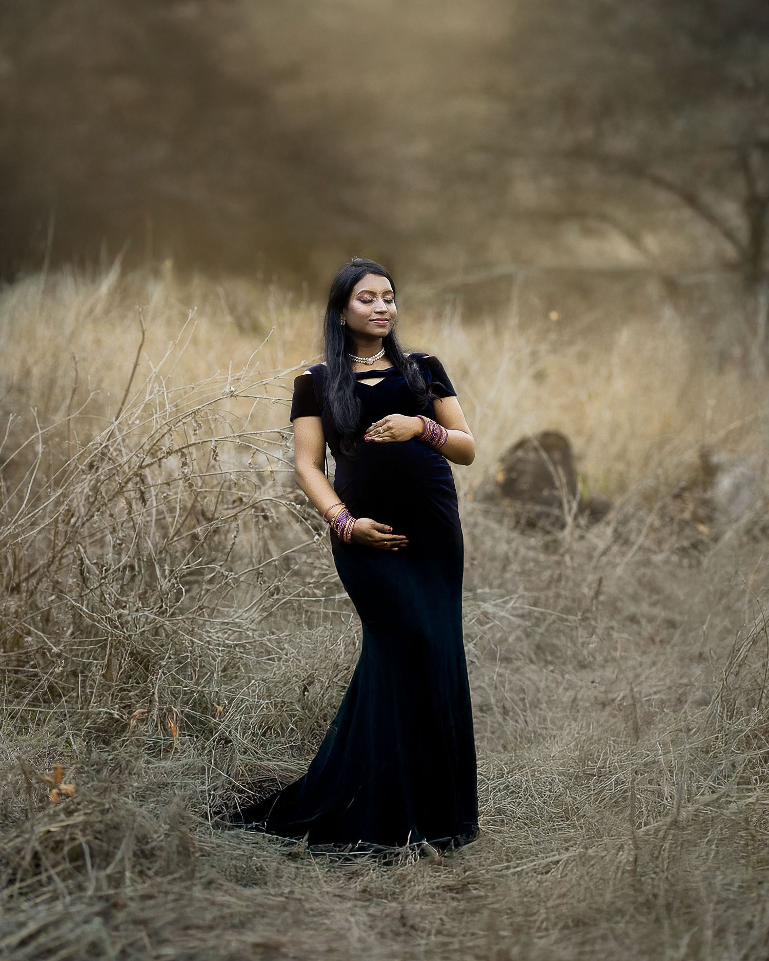 A woman in a black maternity gown poses gracefully in a field of tall golden grass during sunset.