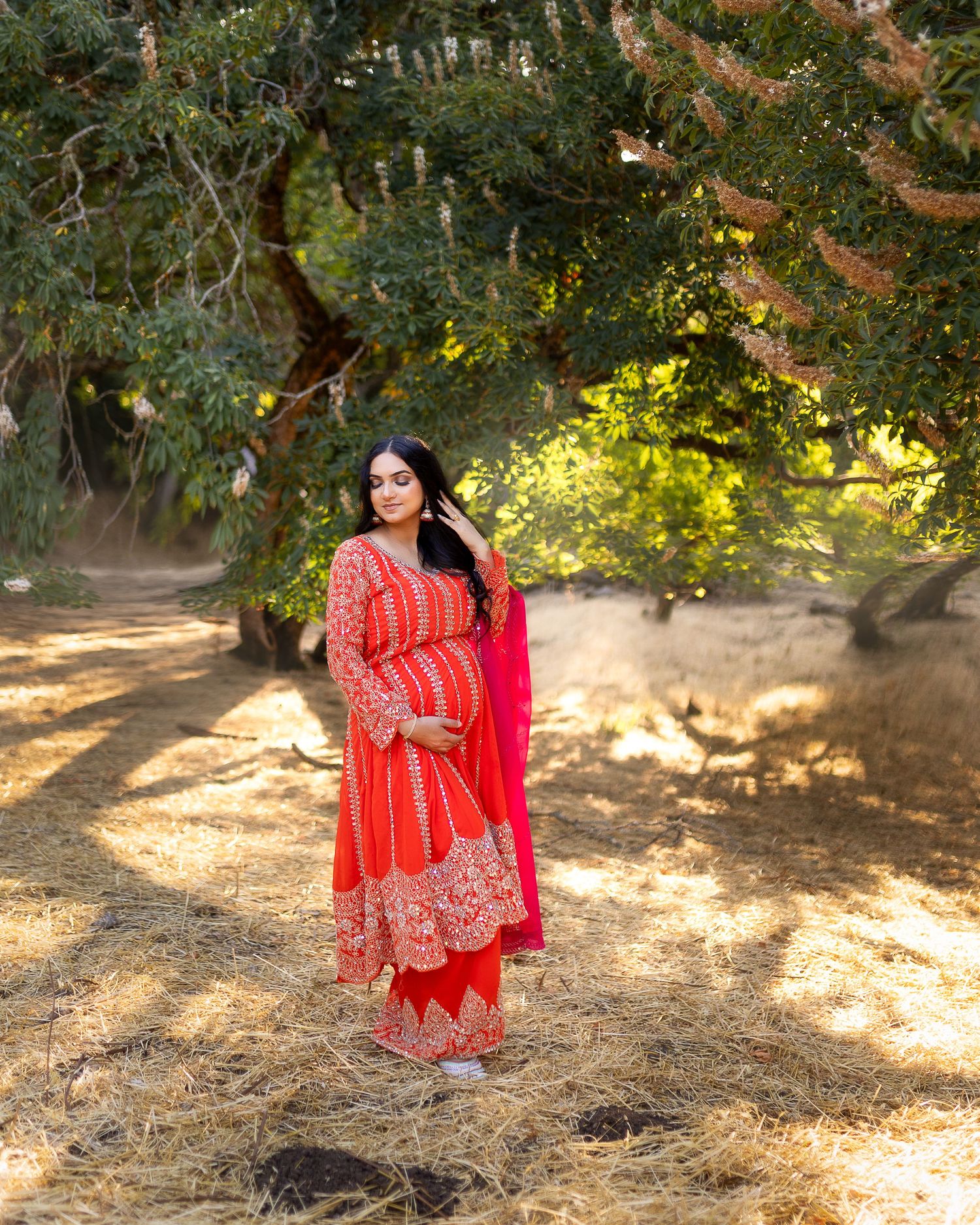 A person in a flowing red and gold traditional Indian dress poses gracefully in a sunlit outdoor setting with trees.
