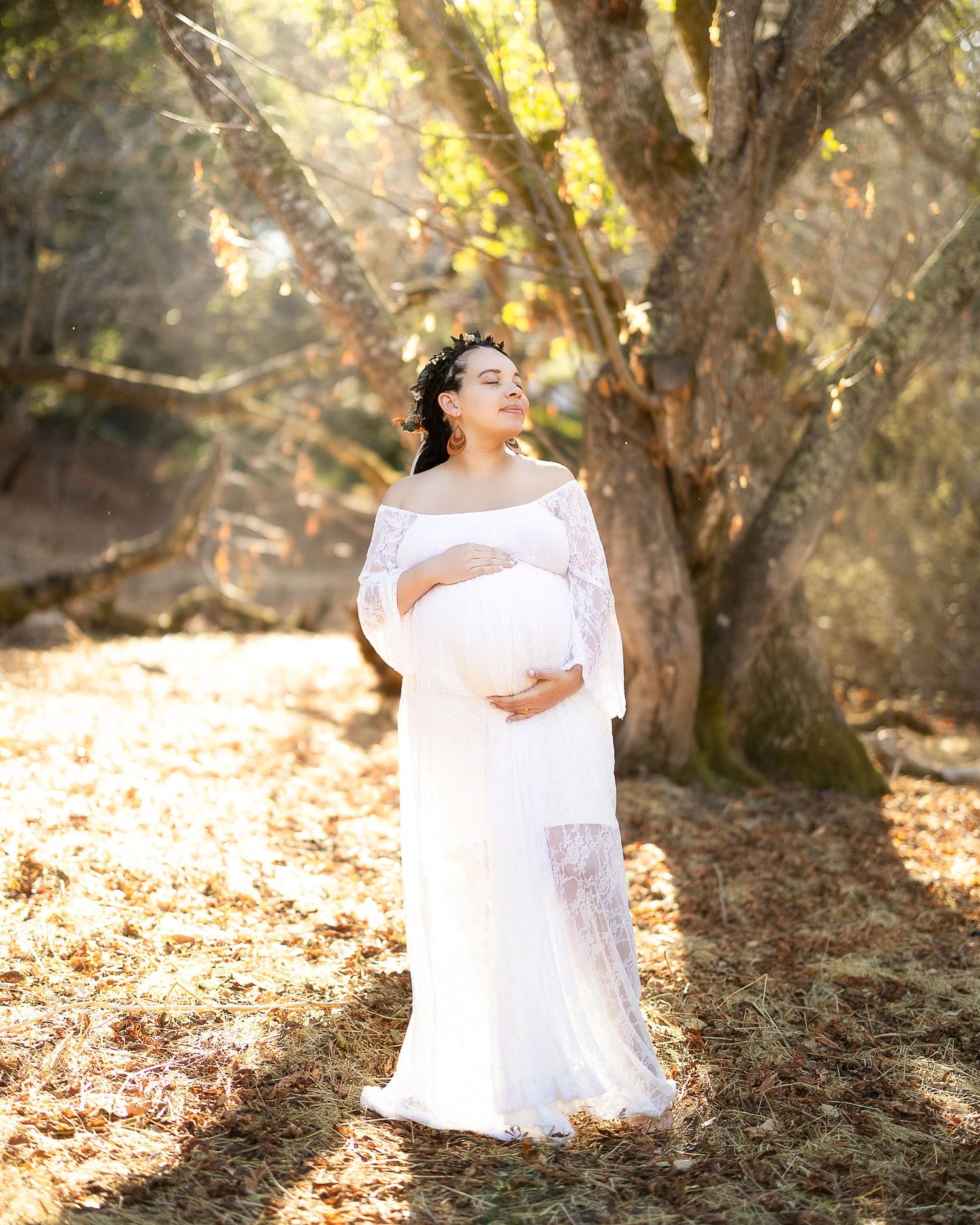 A figure in a flowing white dress poses gracefully among sun-dappled trees in a dreamy outdoor setting.