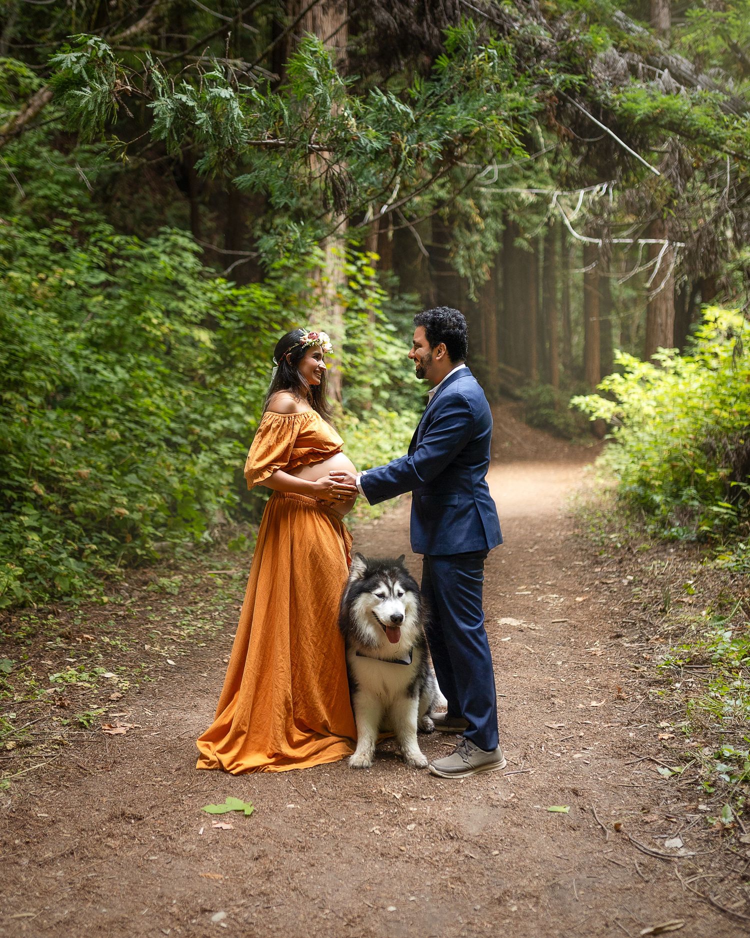 Couple in formal attire poses on forest path with their Husky dog between them.