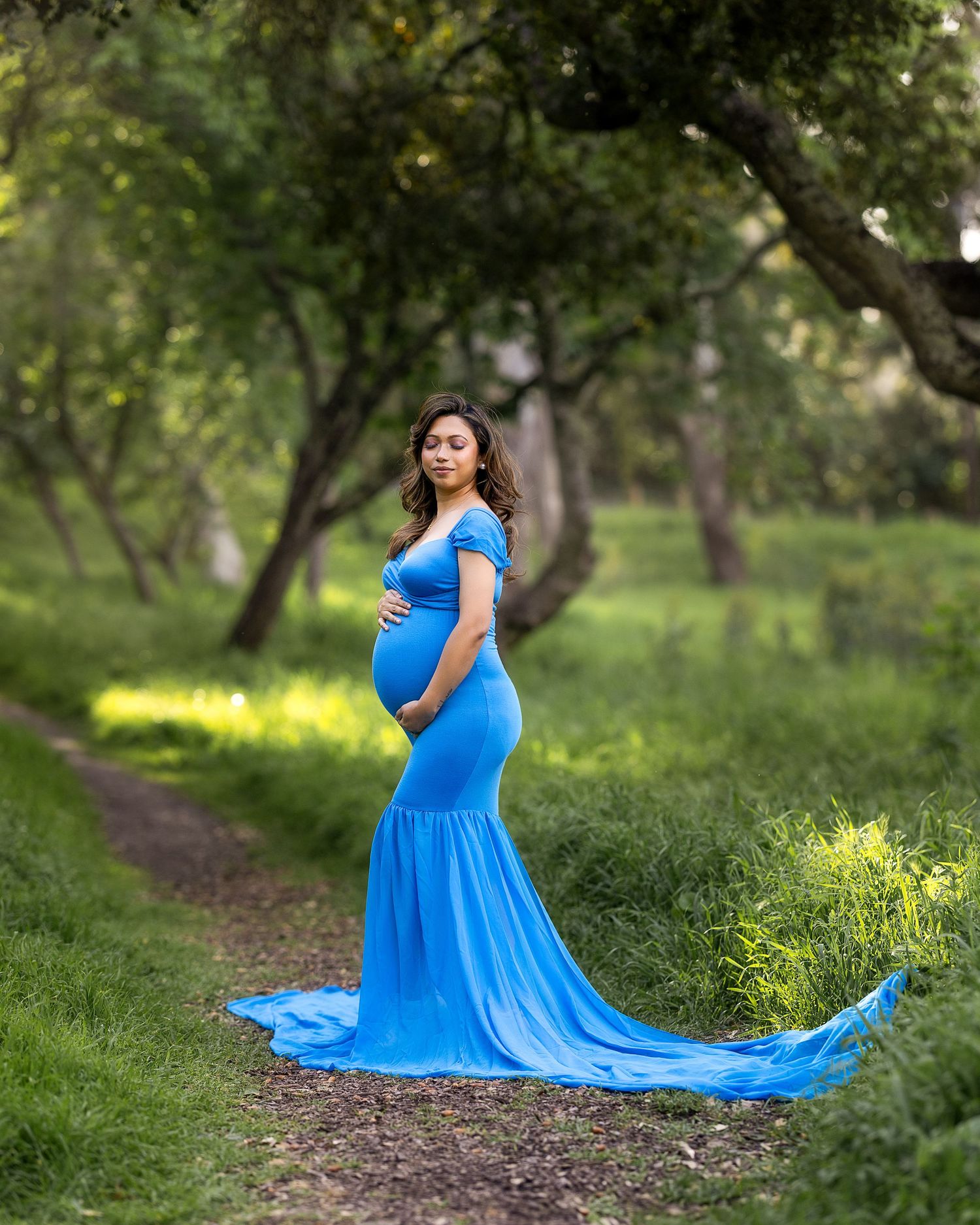 A flowing bright blue maternity gown billows elegantly in a sunlit wooded path during a photo session.