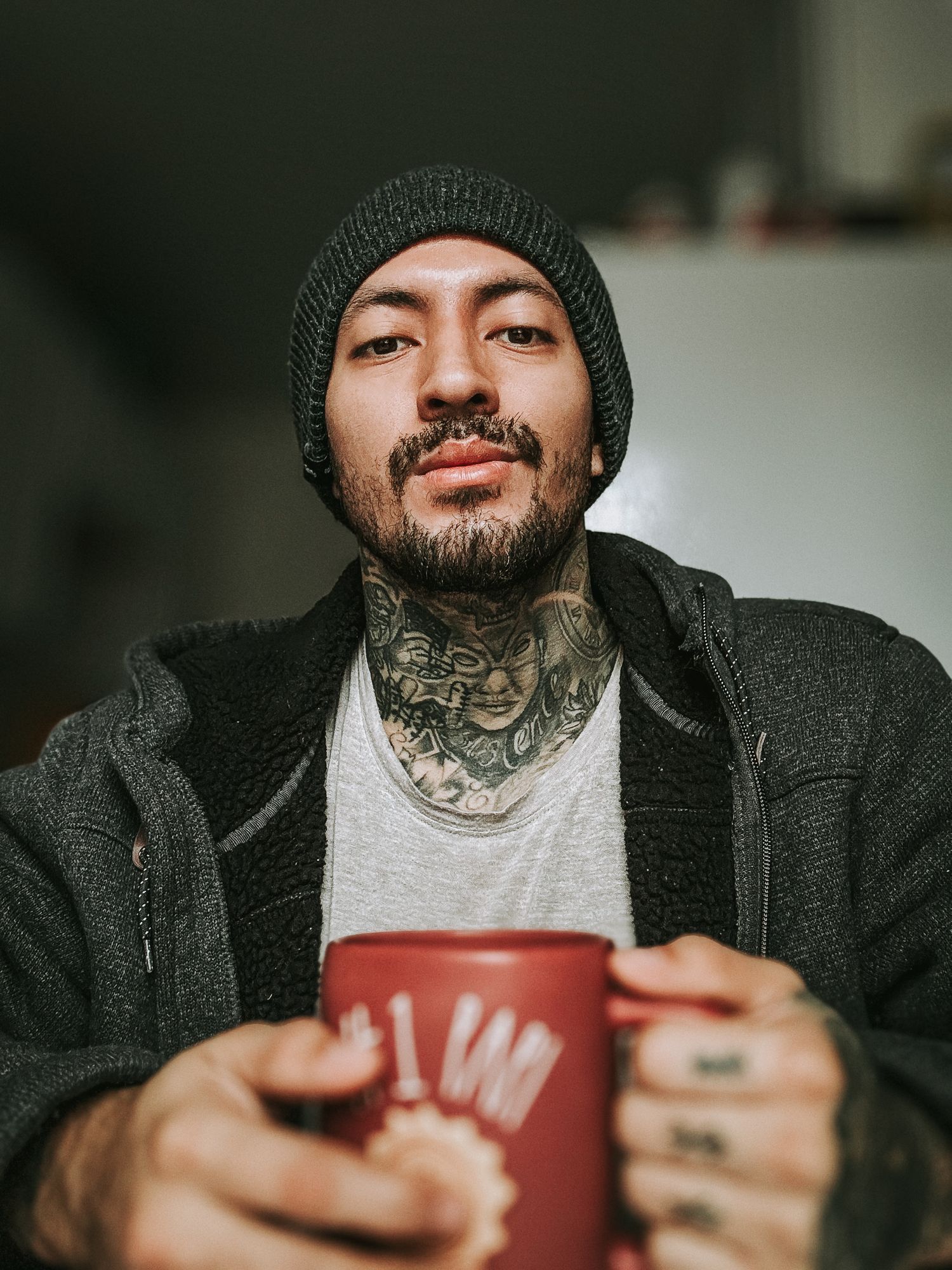 Person wearing dark jacket holds red coffee cup while smiling in moody indoor lighting.