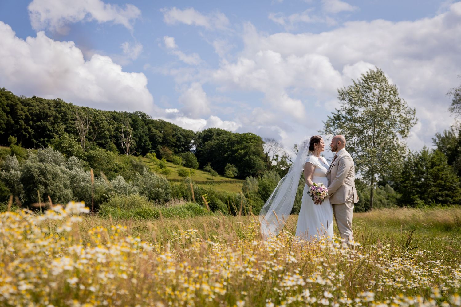 Newlyweds standing in sunny wildflower meadow with dramatic cloudy sky backdrop.