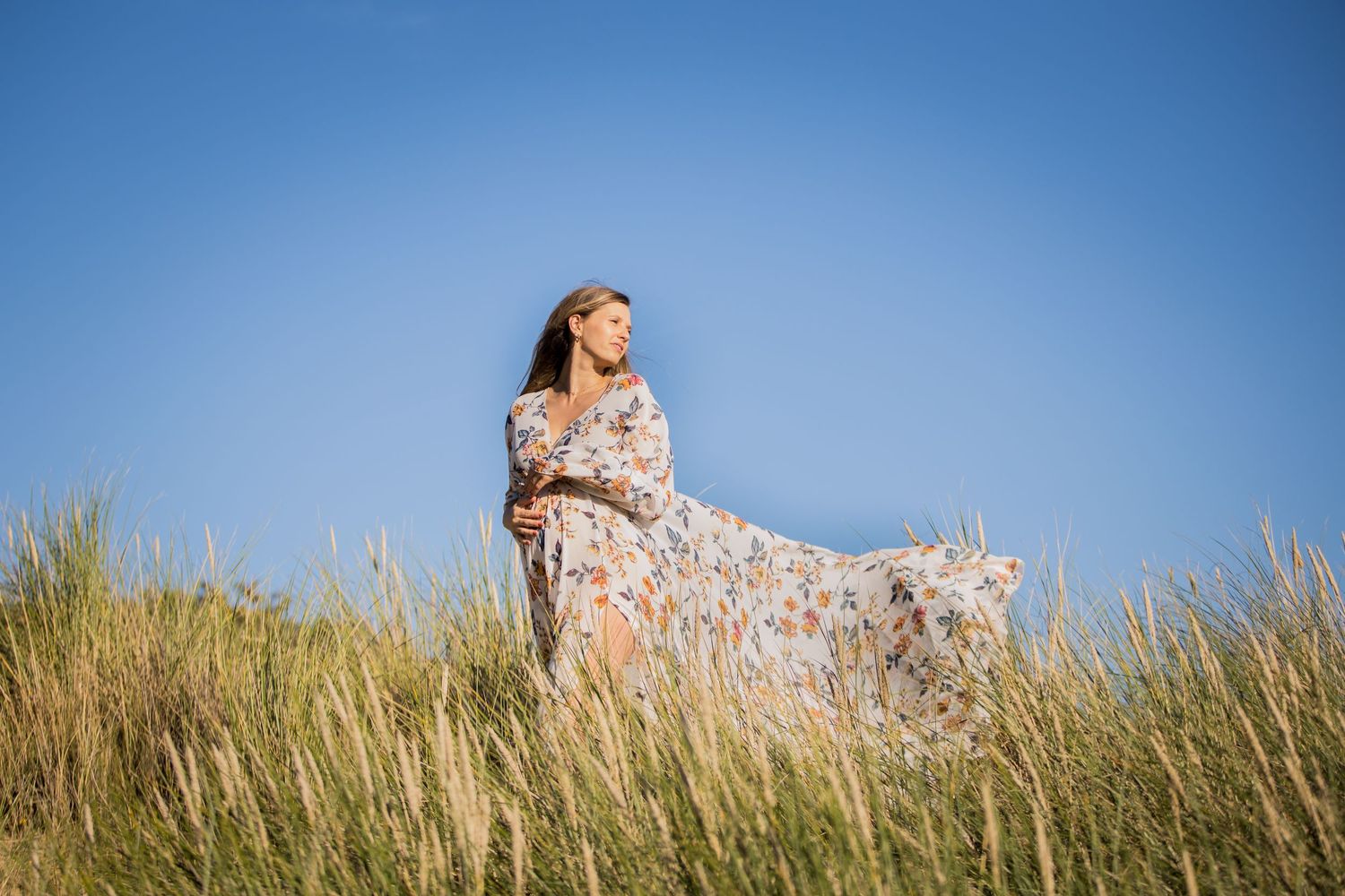 Figure in flowing floral dress stands in tall grass against blue sky.