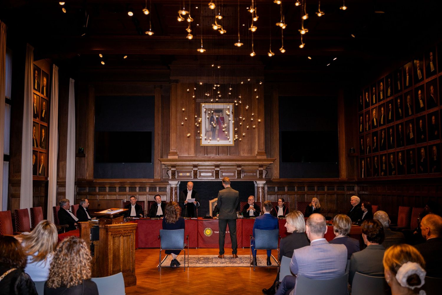 Ceremonial proceedings in an ornate hall with glowing ceiling lights and historic portrait decorations.