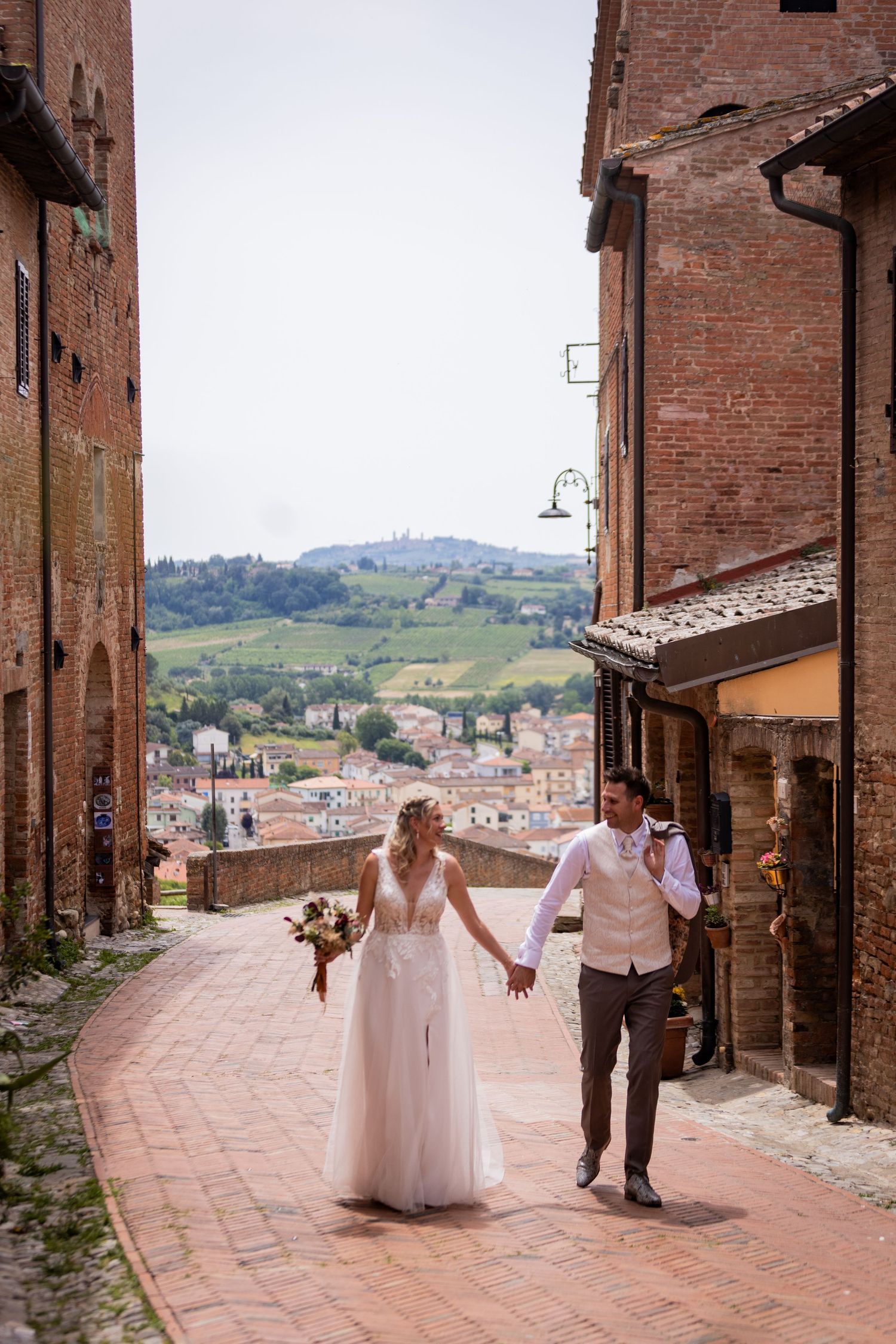 A couple in wedding attire walk down a charming brick street overlooking the rolling hills and red-roofed buildings of Tuscany.