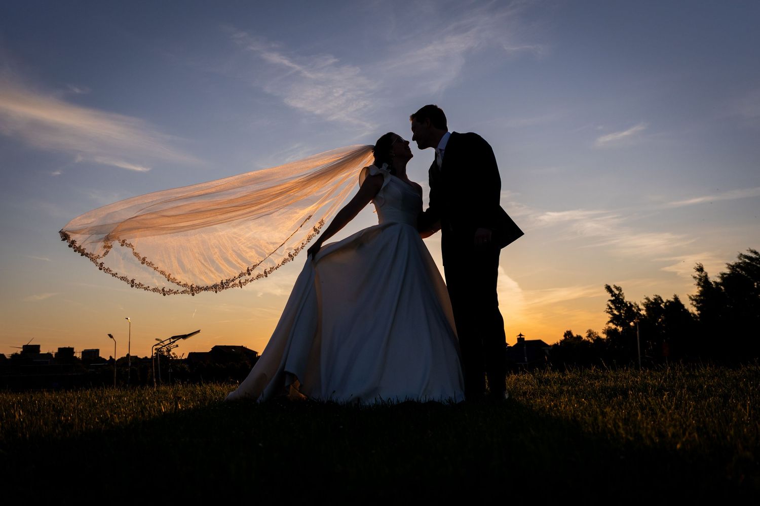 Silhouetted couple in wedding attire share a romantic moment at sunset with flowy veil catching the golden light.