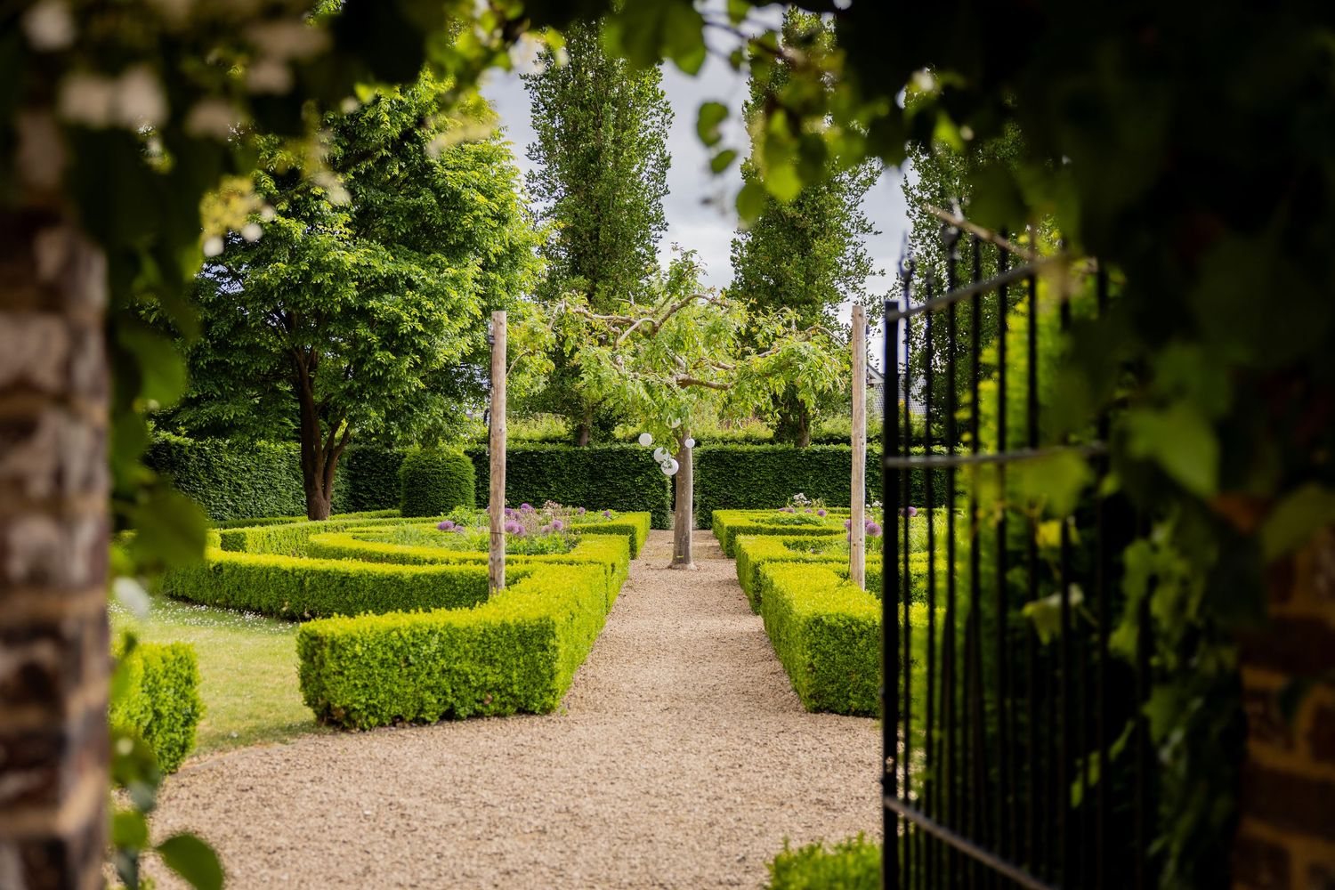 A lush garden pathway with ornate iron gates and manicured hedges leads through a tranquil green parkland setting.