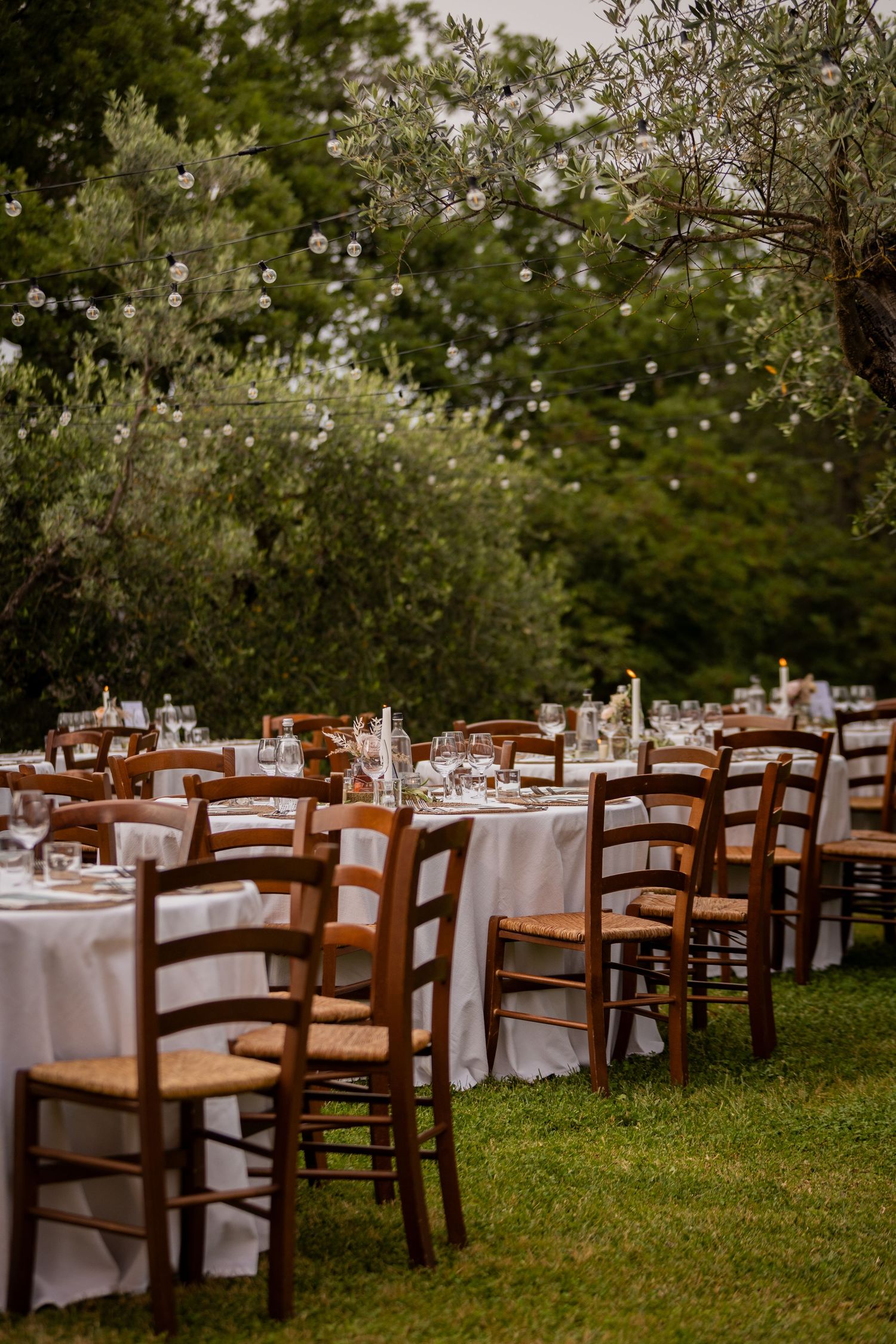 Rustic outdoor wedding reception setup with wooden chairs and white-clothed tables arranged on a grassy lawn under trees.