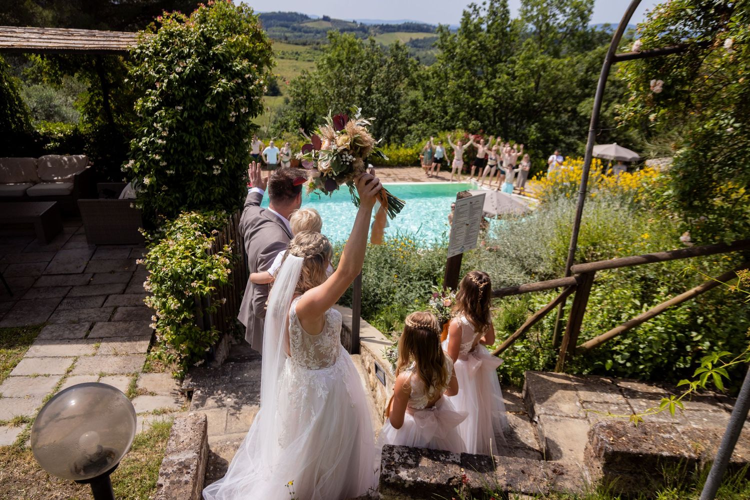 Group in formal attire descend stone steps at outdoor garden venue with pool and mountain views in background.