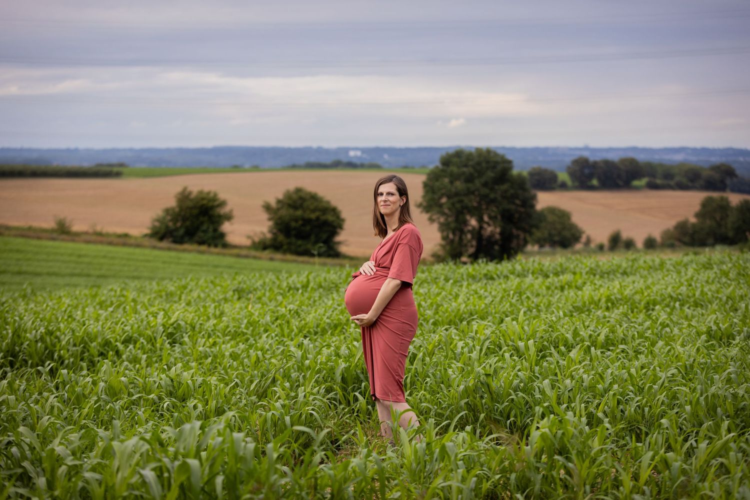 A pregnant woman in a pink dress stands in a lush green field with rolling hills in the background.
