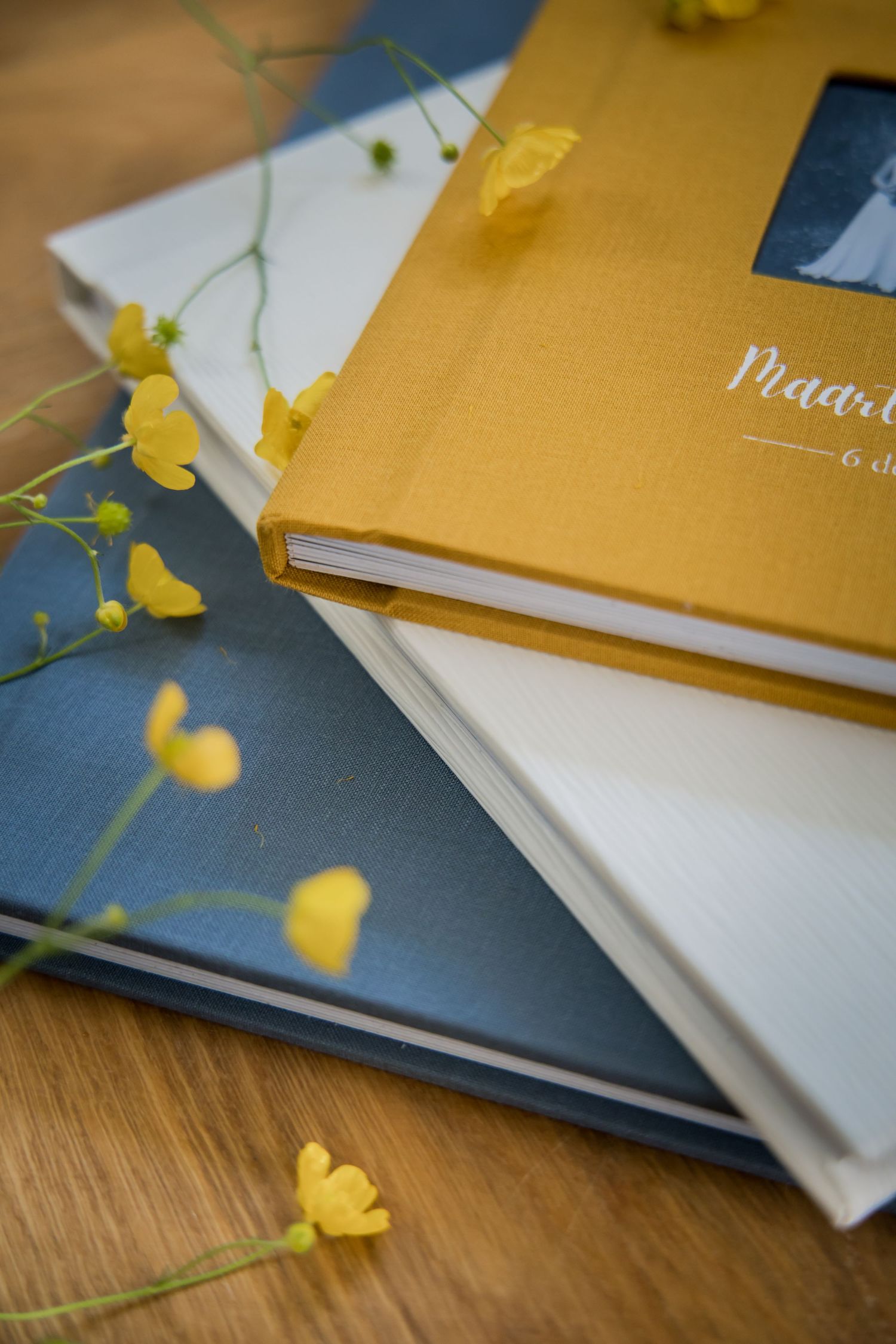 Stacked photo albums or books in gold and navy blue tones with delicate yellow buttercup flowers on wooden surface.