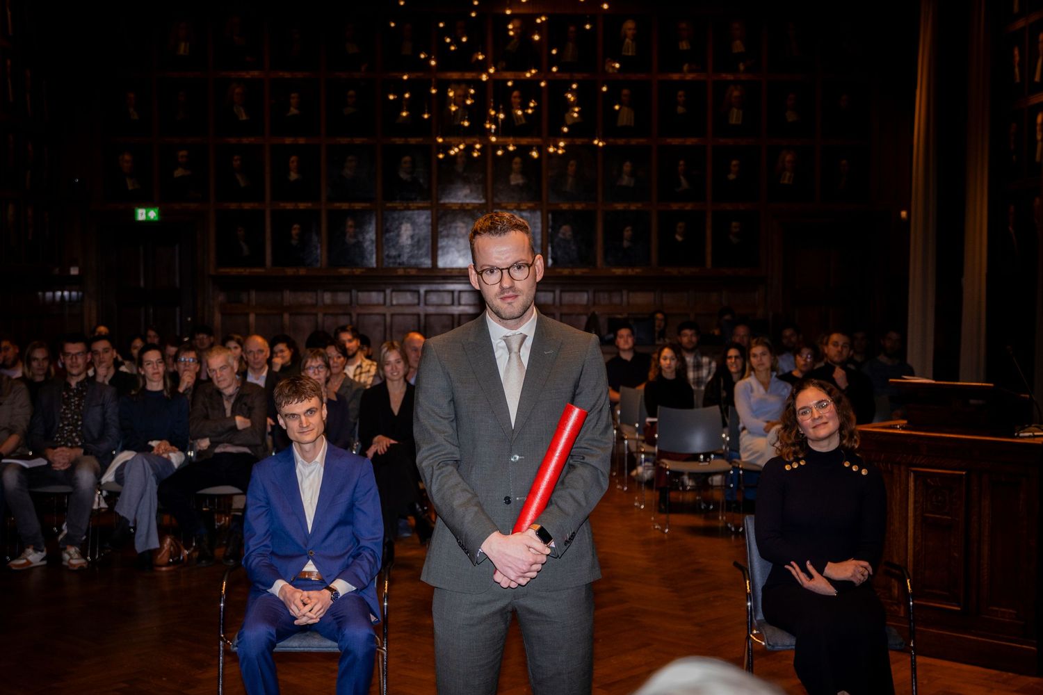 Academic ceremony audience gathered beneath twinkling lights in a historic hall with portrait-lined walls.