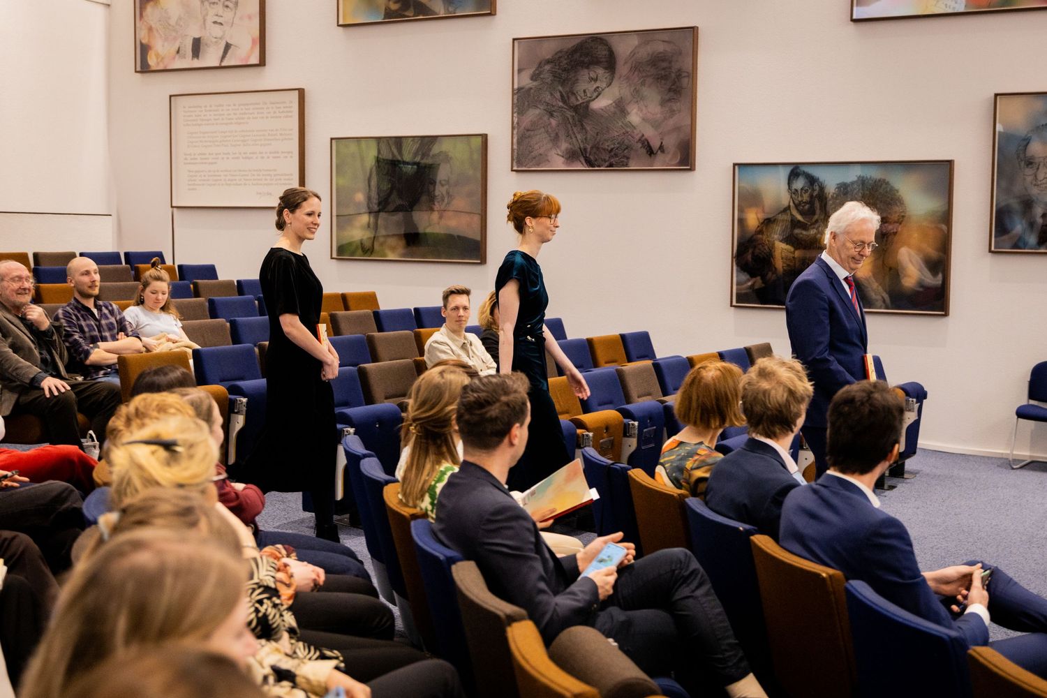 Students and presenters gather in lecture hall with artwork displayed on walls.