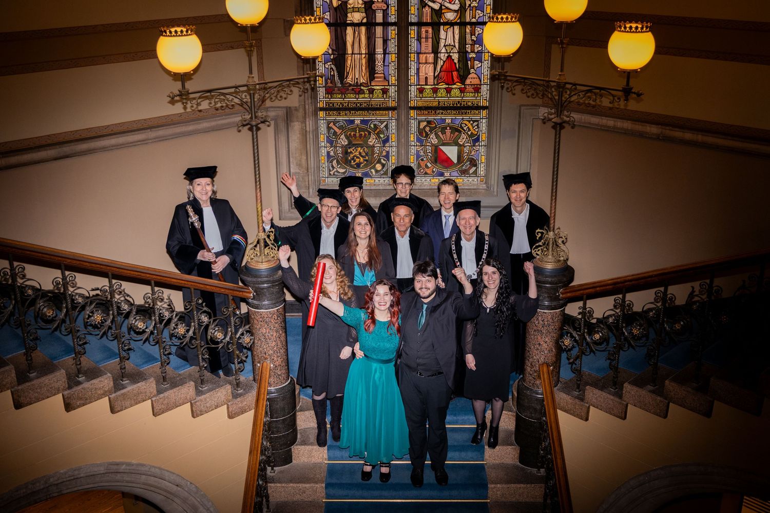 Formal group standing on an elegant staircase with ornate railings and vintage pendant lights.