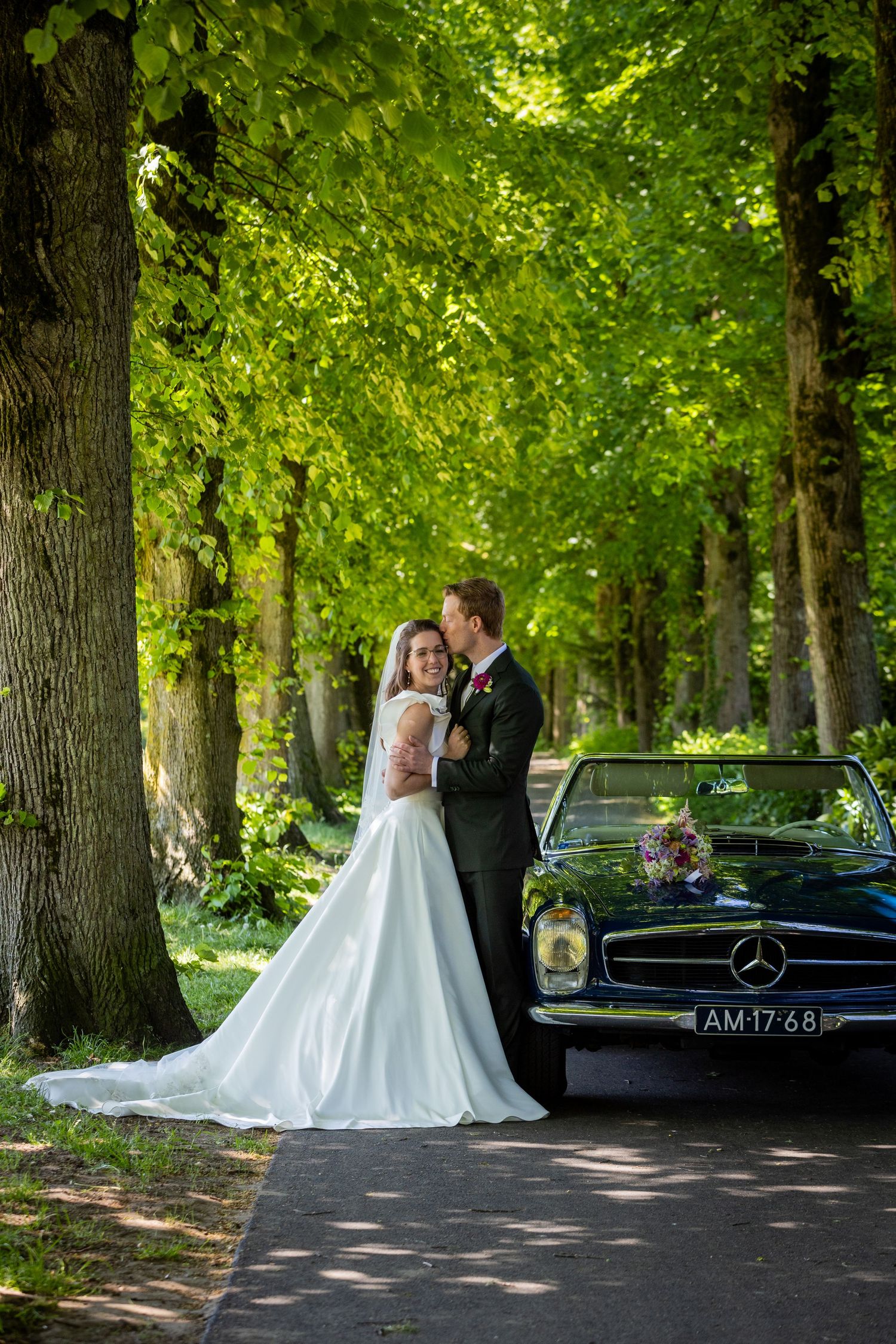 Wedding couple poses with vintage Mercedes-Benz under a canopy of green trees.