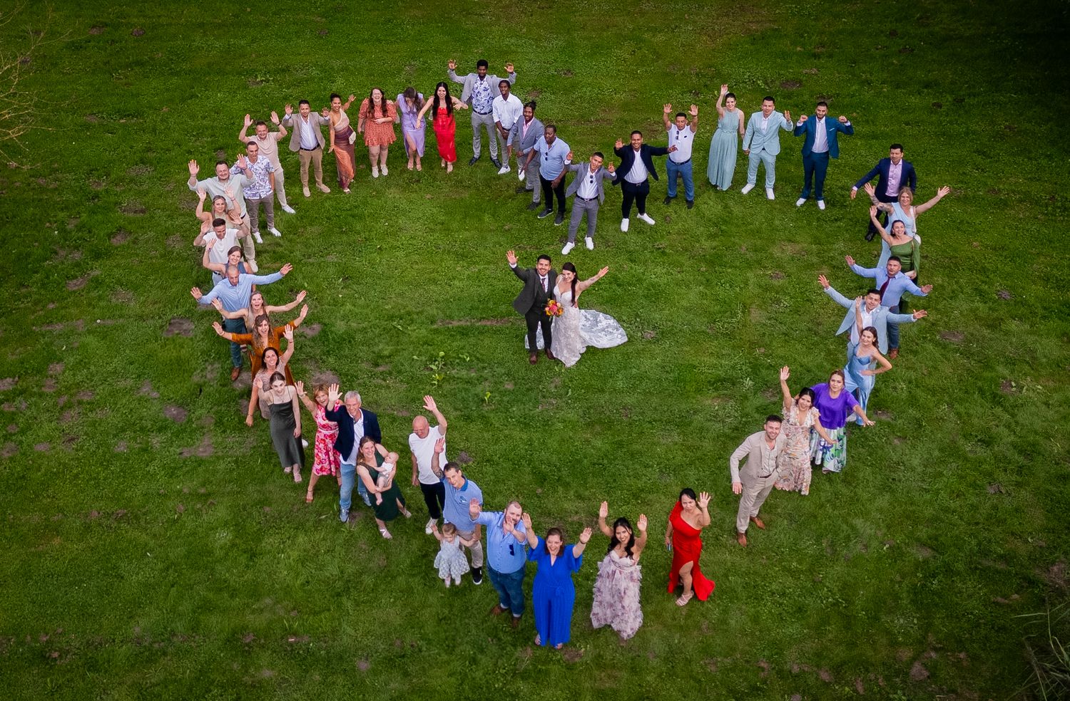 Aerial view of wedding guests forming a heart shape on green lawn.
