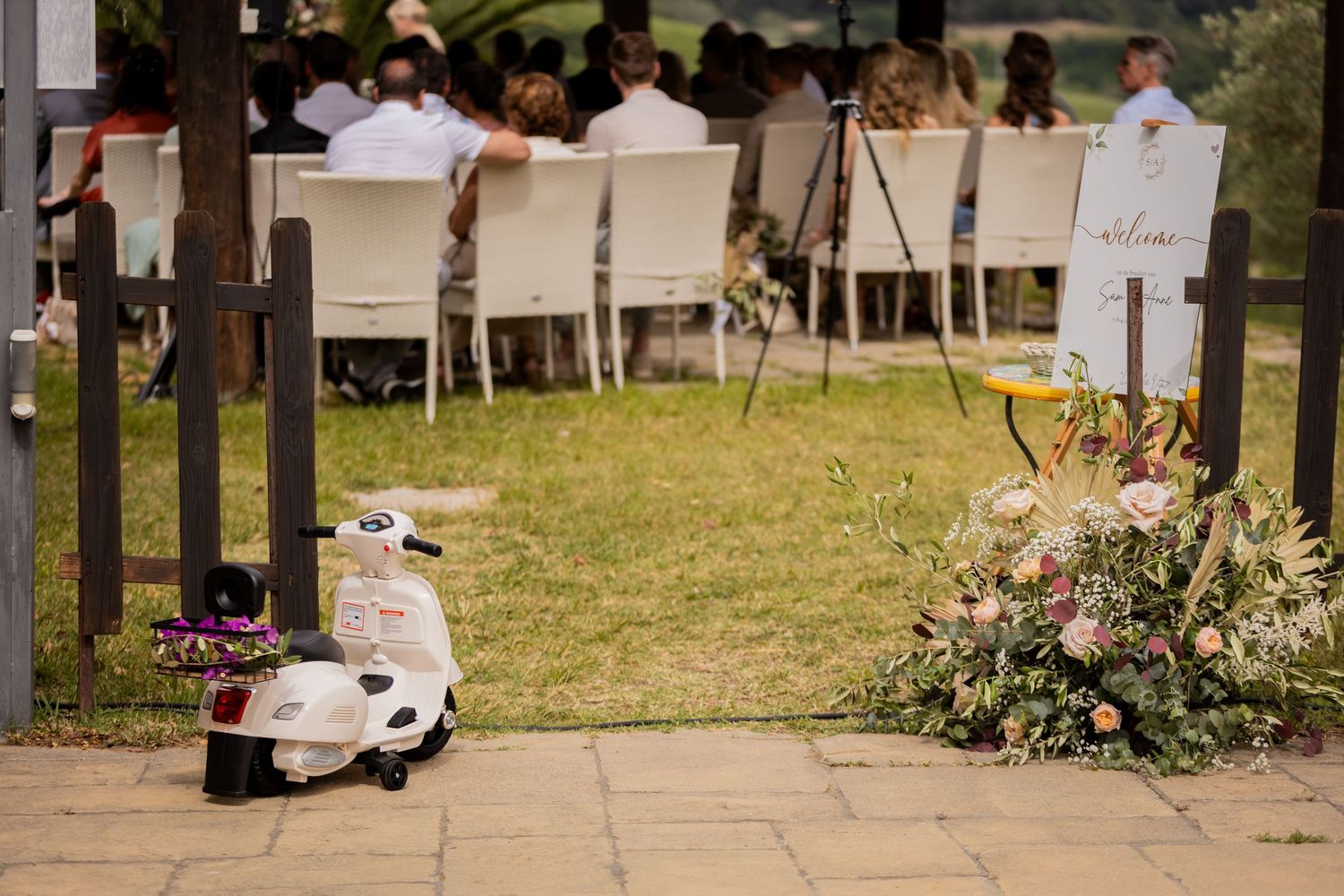 A row of white covered outdoor booths with flowers and people at a market or fair on a sunny day.
