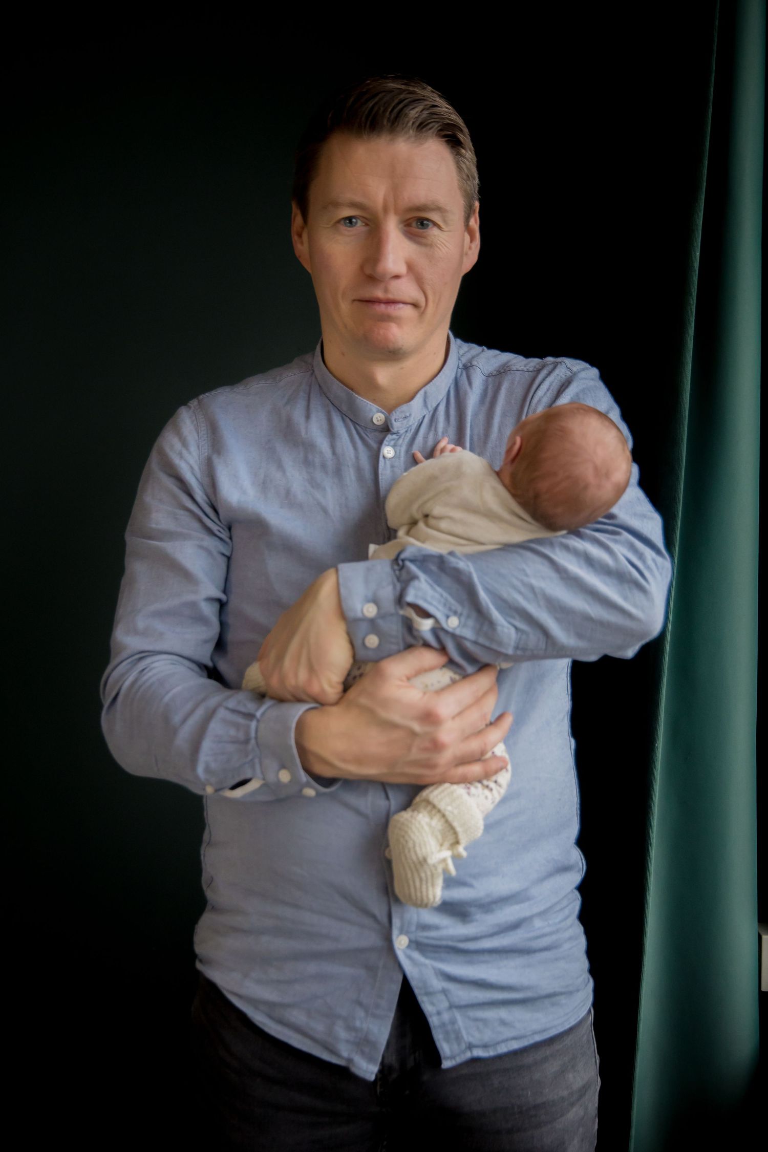Person in blue shirt holding sleeping newborn baby against dark background.