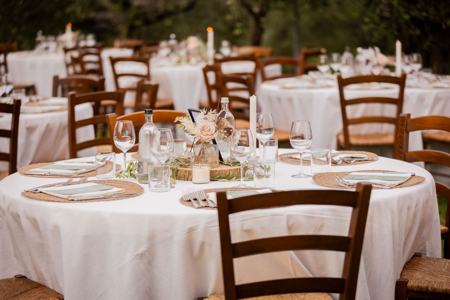 Elegant outdoor dining setup with round tables dressed in white linens and dark wooden chairs for a rustic wedding reception.