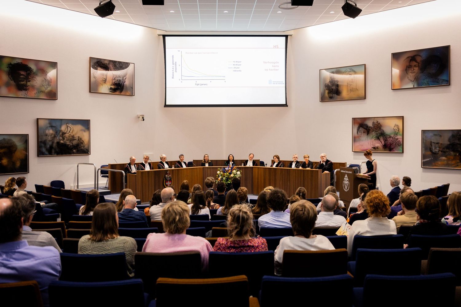 Conference hall with tiered seating filled with attendees facing presentation screen.