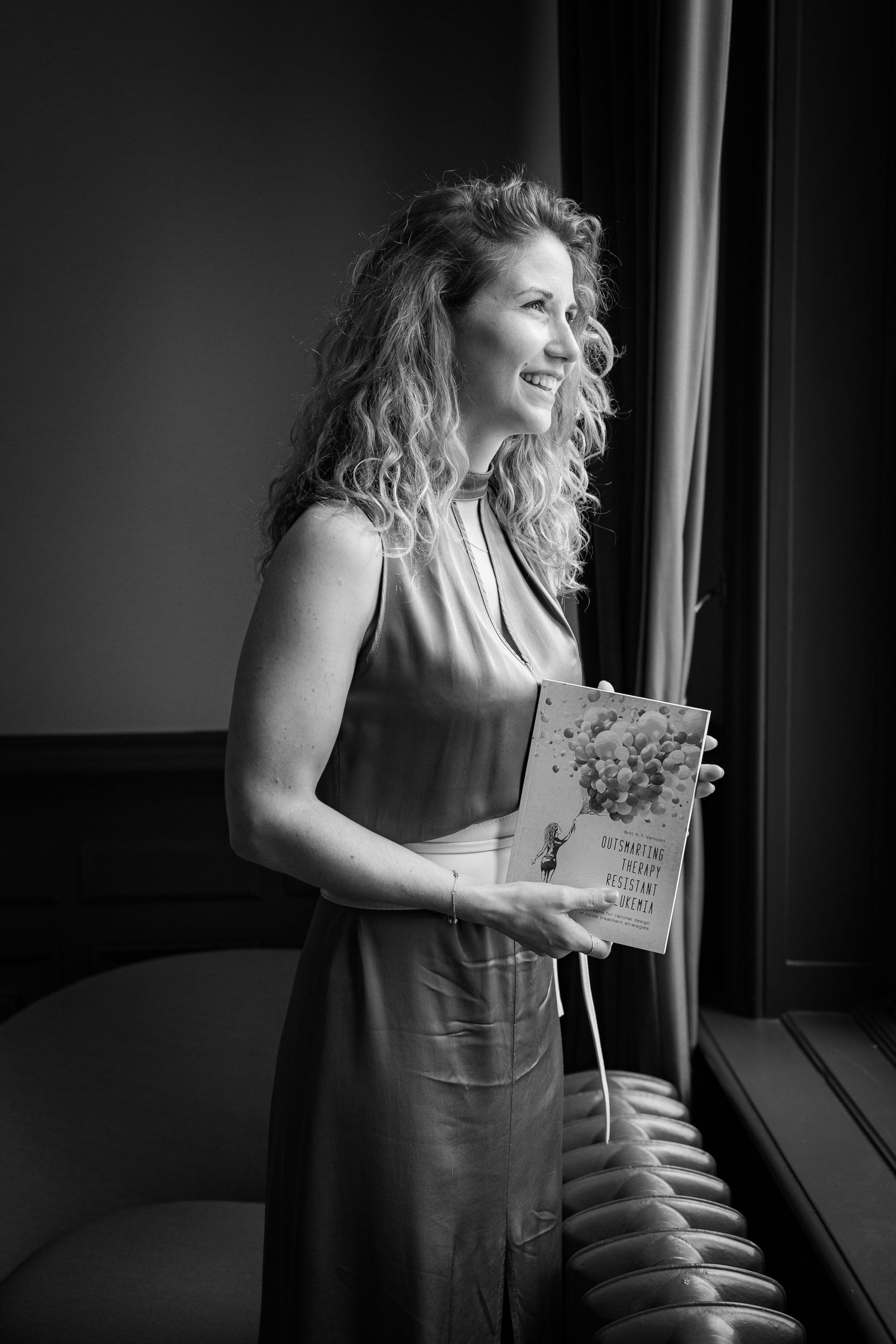 Black and white portrait of person holding a book by window in elegant dress.