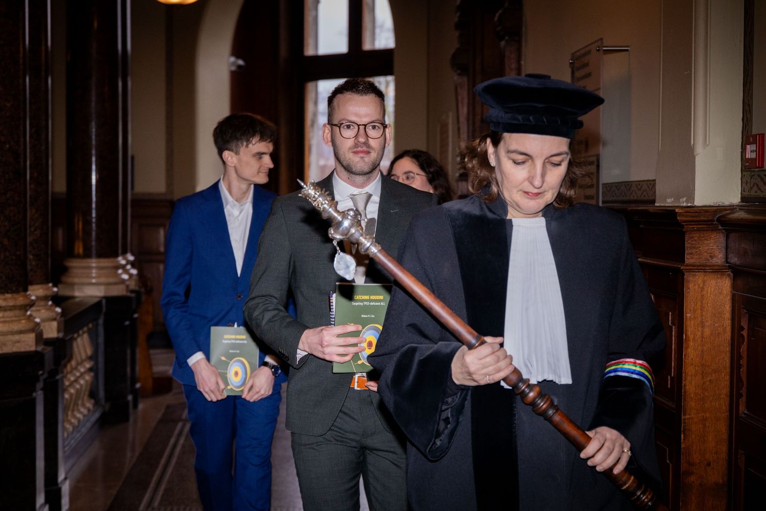 Academic figures walking through a traditional university corridor carrying ceremonial items.