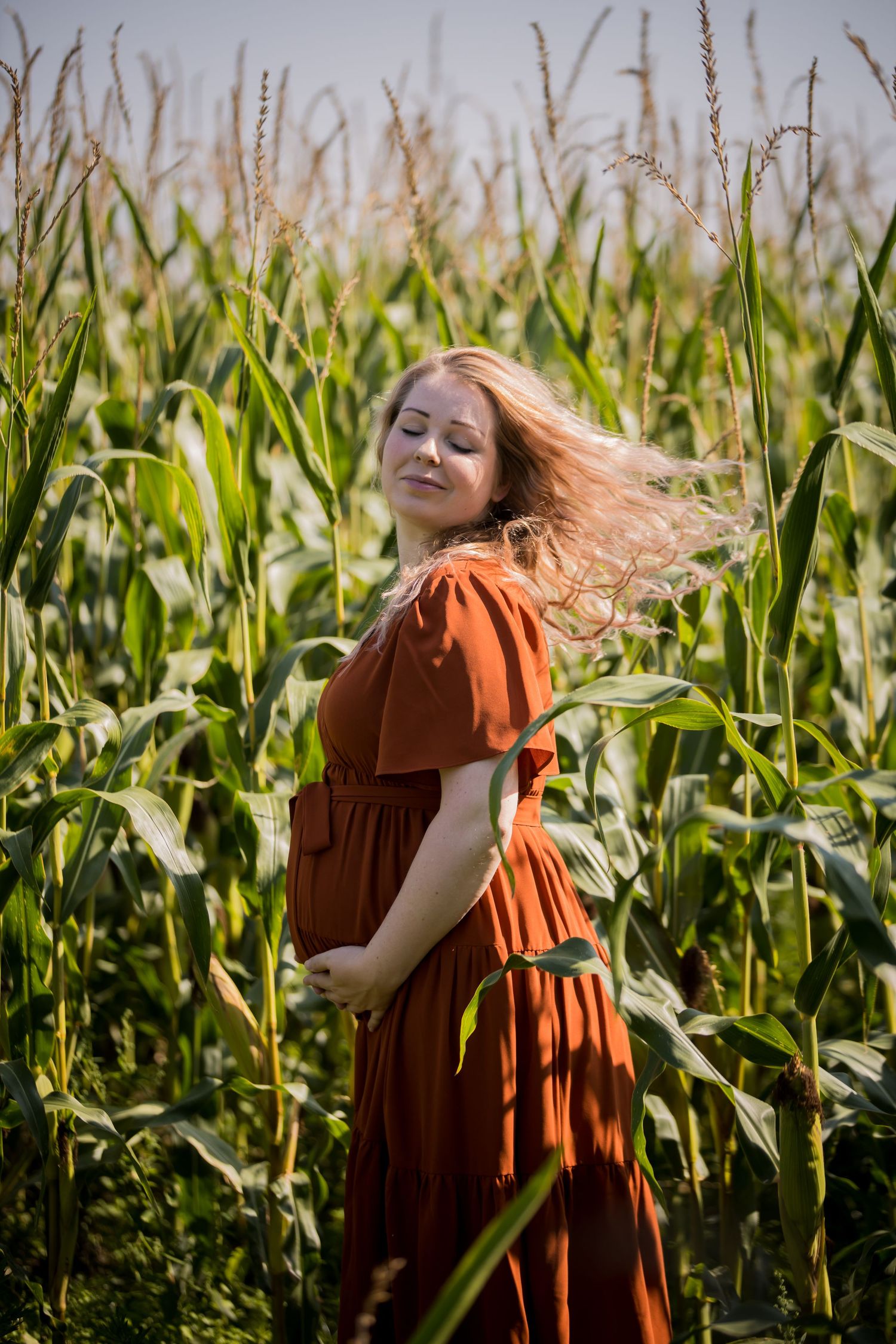 Woman in rust-colored dress twirling joyfully in a sunlit cornfield.