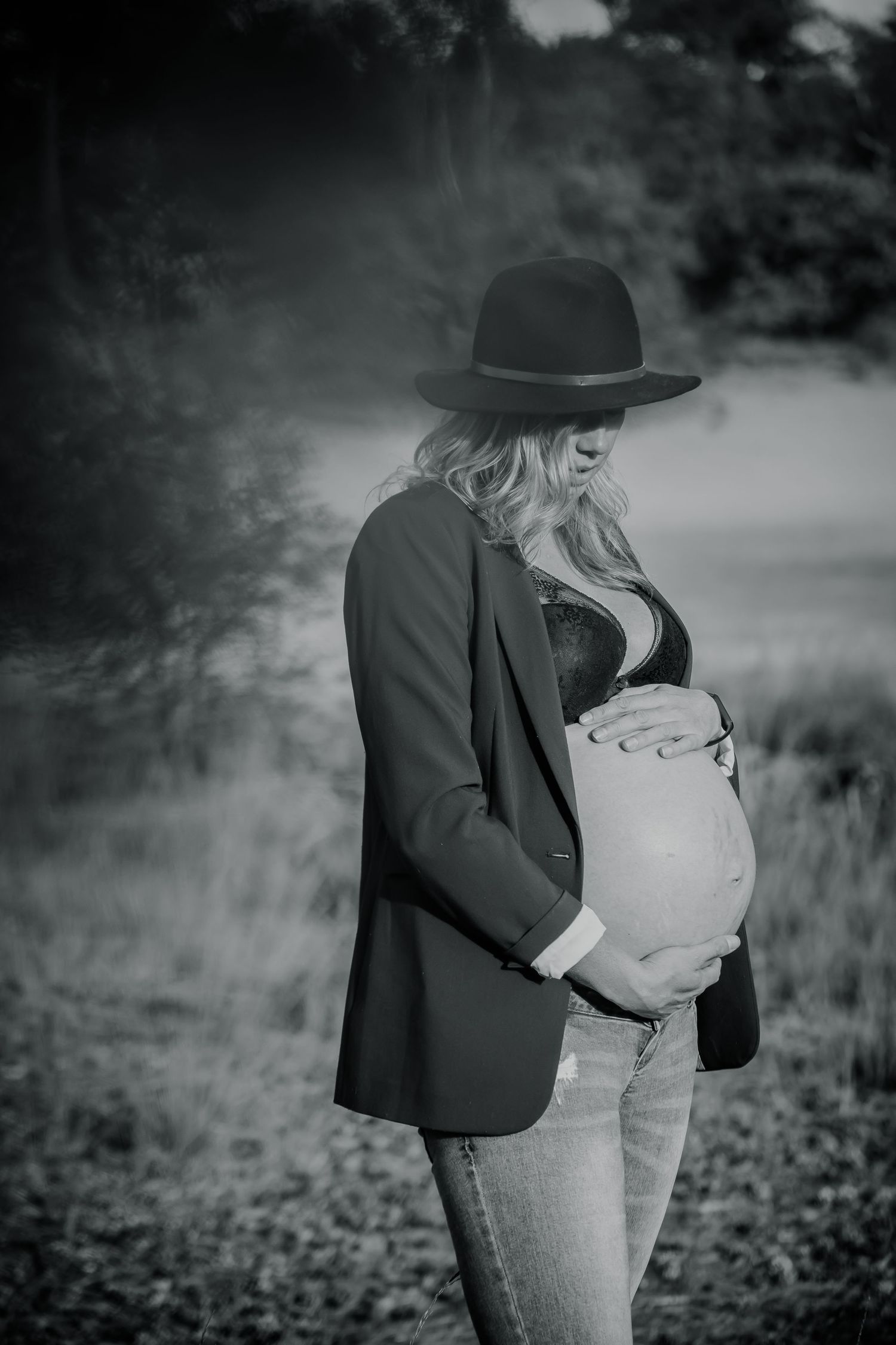 A stylish pregnant woman in a black hat and blazer poses in a moody outdoor setting.