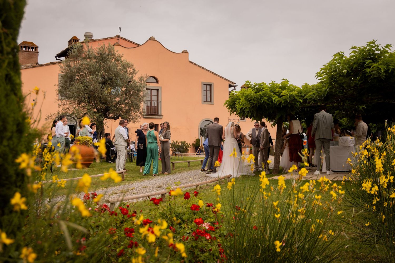 Guests gather outside a peach-colored Mediterranean villa for an outdoor celebration with yellow and red wildflowers in foreground.